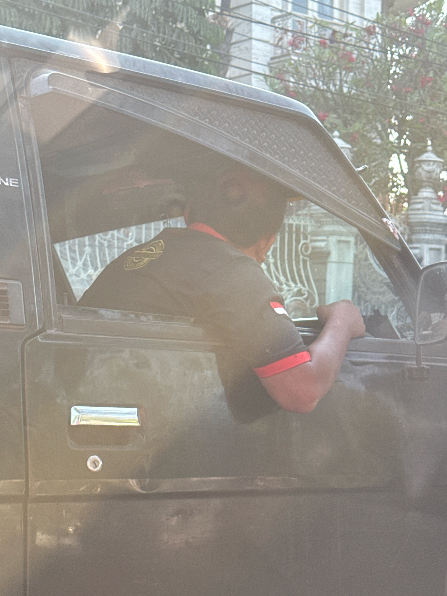 A person is seen through the driver's side window of a dark-colored vehicle, appearing to be in motion or parked. The individual, with dark hair and wearing a dark t-shirt with red trim on the collar and sleeves, has their back partially to the camera. A small Indonesian flag emblem is visible on the right sleeve of their t-shirt. Their left arm rests on the open window frame. The vehicle's interior is dark and indistinct. Outside the vehicle, a residential building with a light-colored facade and ornate architectural details is visible, along with lush greenery and flowering trees. The scene appears to be captured during daylight, with a slight lens flare in the upper left quadrant of the image. The presence of the Indonesian flag emblem suggests a possible location within Indonesia, and the architectural style of the building may be indicative of specific regions or cities within the country. Based on common architectural styles and vegetation observed in the image, this scene could potentially be located in a city like Surabaya, Indonesia, which features a blend of traditional and modern residential architecture and abundant tropical flora.