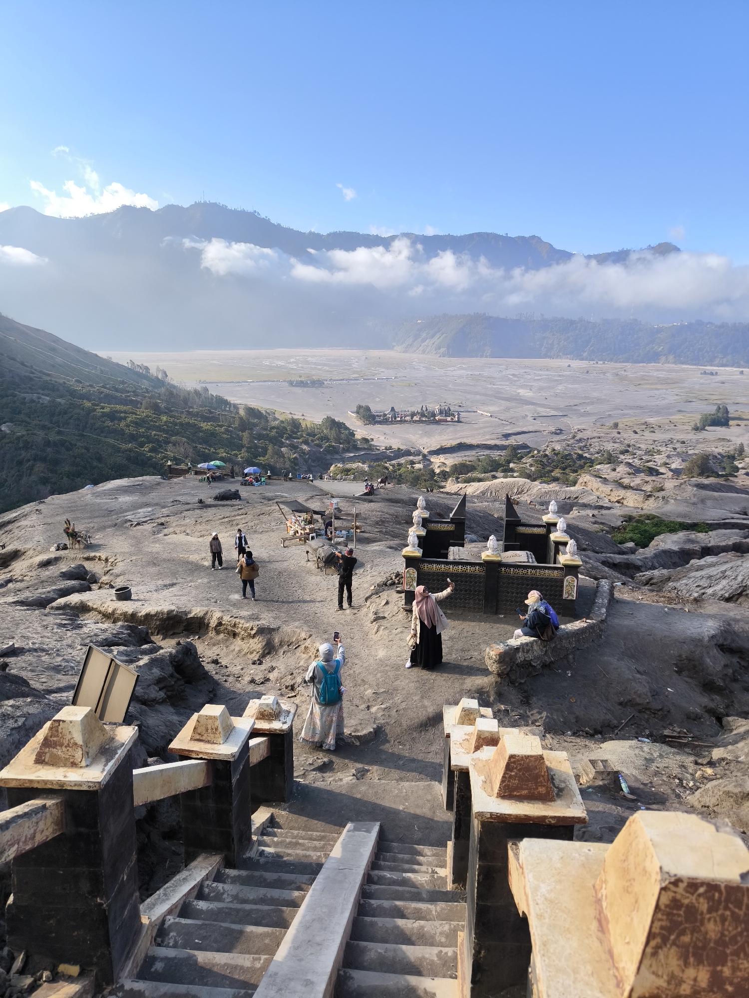 This image captures a scene at Cemoro Lawang, Indonesia, likely at sunrise or sunset due to the warm, soft lighting and long shadows. The sky is a clear blue with some scattered clouds, and the distant mountains are partially veiled in mist.
The foreground features a set of stone stairs leading down, flanked by weathered concrete pillars topped with decorative finials. Beyond the stairs, the ground is rocky and uneven, with a small, ornate black structure with white detailing in the mid-ground. This structure appears to be a shrine or monument, with some people gathered around it.
Several individuals are visible in the scene. In the foreground, a person wearing a light-colored headscarf and a blue backpack is taking a photograph with their phone. To the left, another person in a light headscarf and a long black skirt is also taking a photo, possibly a selfie. Further back, a group of people are walking and standing, some appearing to be tourists. There are also some stalls or temporary structures with umbrellas set up in the background, suggesting a tourist area.
The overall setting is a volcanic landscape, with the dry, sandy plains of a caldera stretching into the distance, bordered by rolling hills and distant, rugged mountains. The ground appears to be volcanic ash and sand, with sparse vegetation clinging to the slopes. The mood is one of tranquil exploration and appreciation of the natural scenery. There is no visible text in the image.