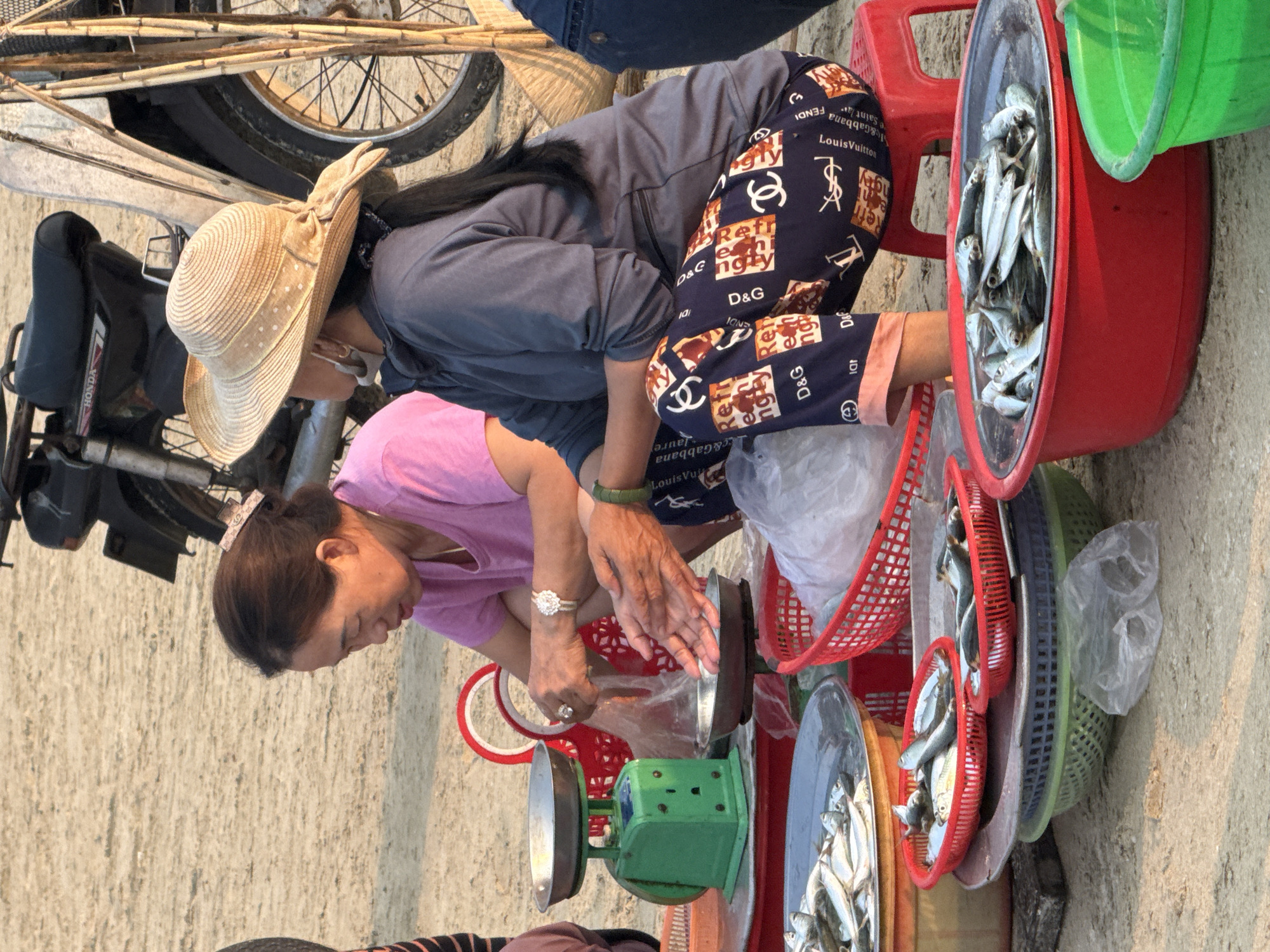 The image shows two women weighing fish on a scale on the streets of Đà Nẵng, Vietnam. The woman in pink is operating the scale, while the woman in gray is assisting. Several baskets filled with fish are on display. A motorbike is parked nearby. The setting appears to be an outdoor market or roadside stall. The scale is green with a metal pan.
