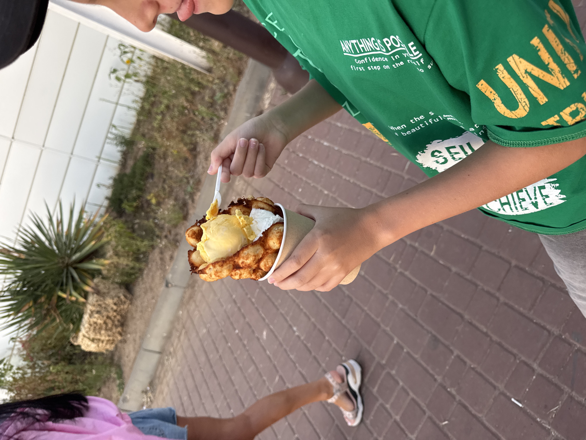 In Novofedorovka, Ukraine, a person is holding a bubble waffle cone filled with ice cream and whipped cream, with a plastic spoon in their hand. The person is wearing a green t-shirt with text. Another person, wearing a pink shirt and denim shorts, walks away in the background on a brick pathway. The environment includes dry vegetation and a white building.