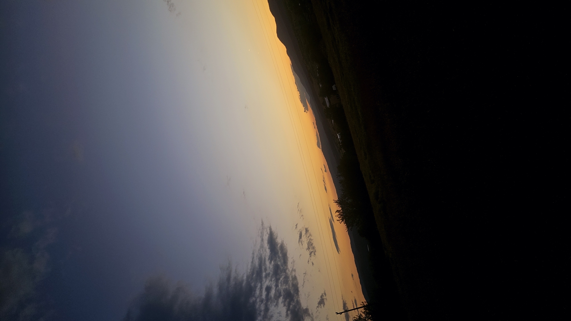 The image shows a sunset over a hilly landscape. The sky transitions from dark blue at the top to a warm orange and yellow near the horizon. Silhouetted against the sky are rolling hills, trees, and what appear to be a few scattered buildings. Power lines are visible running diagonally across the frame. The foreground is dark and indistinct, obscuring any details of the ground. The scene appears to be in a rural or semi-rural area.