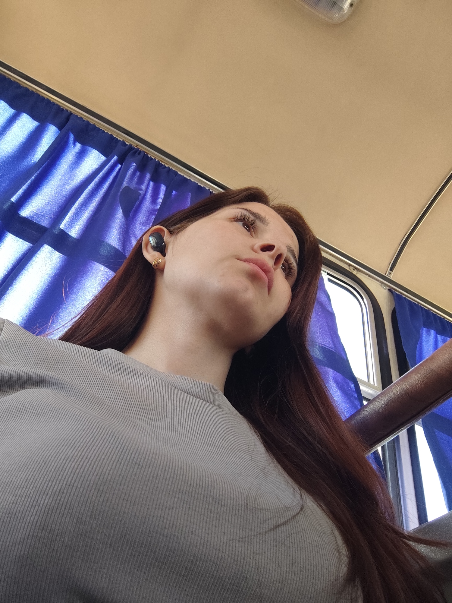 A woman with long auburn hair, wearing a gray ribbed shirt and gold earrings, is seen from a low angle. She has an earbud in her left ear and is looking upward. She appears to be on a bus or a similar vehicle, as indicated by the blue curtains, the window frame, and the overhead lighting. The interior ceiling of the bus is tan. There is a brown handrail visible. The location is unknown and cannot be determined from the information provided in the image.