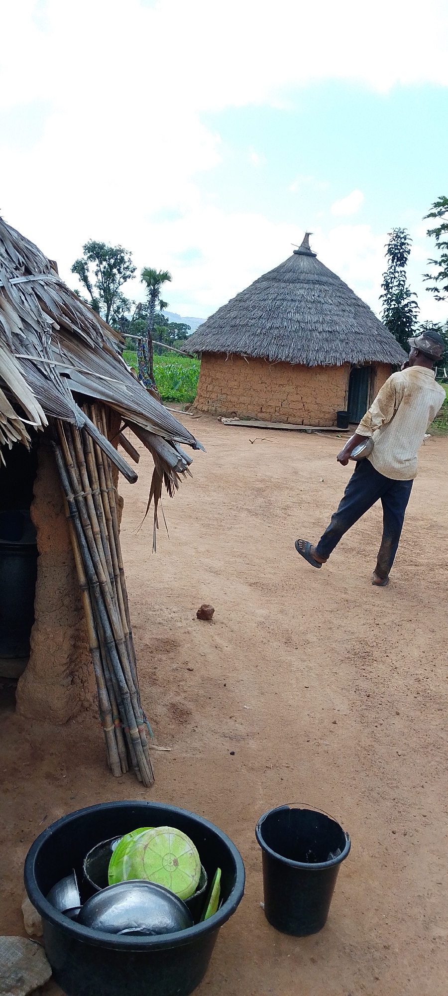 A man is walking away from the camera in a dusty, rural setting. He is wearing a light-colored, long-sleeved shirt and dark pants, with a cap on his head. He is holding a circular object in his right hand. The ground is dirt and sand, with some scattered small stones. In the foreground, to the left, is a black bucket filled with various metal bowls and green circular objects, possibly lids. To the right of this bucket is another black bucket.
Behind the man, there are two traditional thatched-roof huts. The hut on the left is partially visible and appears to be constructed of mud with bamboo poles supporting its roof. The hut on the right is a round, mud-brick structure with a conical thatched roof. In the background, there are some trees and vegetation, with a hint of a cornfield and a distant hilly landscape under a partly cloudy sky. The overall environment suggests a village setting in a warm climate. This scene is located in Kamantan, Nigeria.