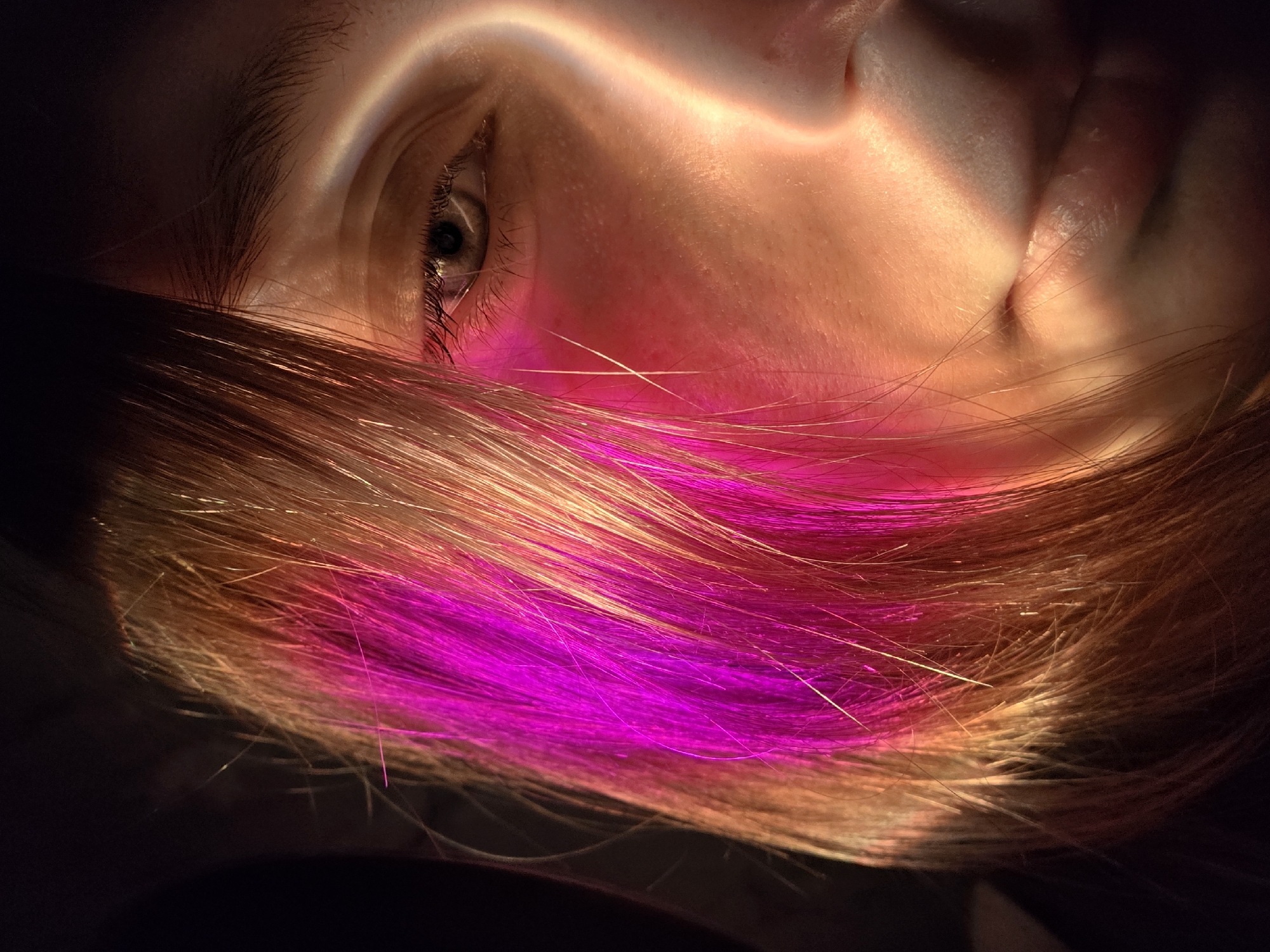 The image depicts a close-up, vertical portrait of the right side of an individual's face, from the brow to the upper lip. A prominent vertical band of warm, golden light illuminates the skin, creating a sharp contrast with the shadowed areas. The right eye is open and visible, with a light-colored iris and dark pupil. The bridge and right side of the nose are lit, as is the upper lip and the corner of the mouth, which shows a slight upturn. Strands of hair, a mix of light brown and vivid magenta/fuchsia, frame the left side of the face, some also catching the light. The background is uniformly dark and indistinct, offering no specific environmental context. The location is undetermined, in the city of Unknown, Unknown.