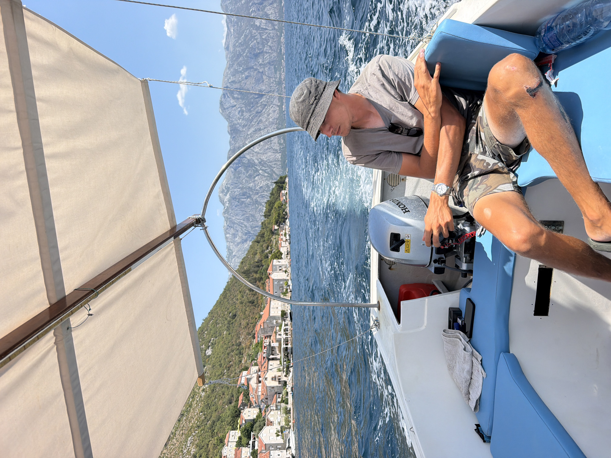 A man wearing a grey t-shirt, camouflage shorts, and a grey bucket hat sits on a white boat with blue seats. He is looking to his left, with his left arm resting on his knee and his right arm resting on the back of the seat. The boat is moving through blue water, with a wake trailing behind it. In the background, there is a town with red-tiled roofs nestled against a green, mountainous hillside. The town is situated along a bay, with more mountains in the distance. The sky is blue with a few white clouds. The scene appears to be a sunny day in Perast, Montenegro.