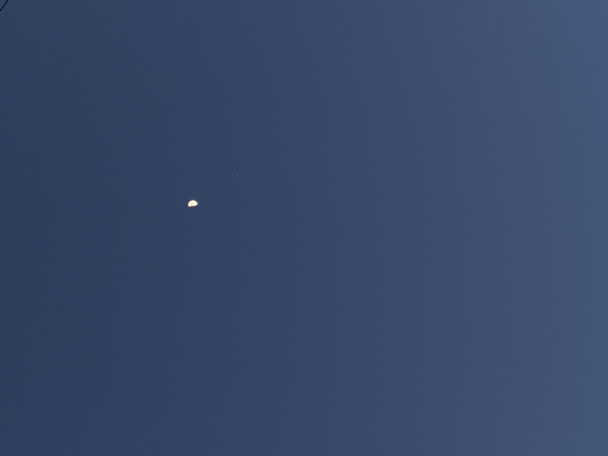 The image displays a dark blue, cloudless sky, indicative of twilight or nighttime conditions. A distinct, white crescent moon is visible in the upper central portion of the frame, illuminated brightly on its right-hand side. Situated approximately one moon-diameter below and slightly to the right of the moon is a faint, minute white point of light, consistent with a star or planet. No other celestial bodies, atmospheric phenomena, ground features, man-made structures, people, or identifiable objects of terrestrial origin are present within the frame. The scene is observed from what has been specified as Perm, Russia; however, the celestial content of the image does not provide visual cues to confirm a specific terrestrial location.
