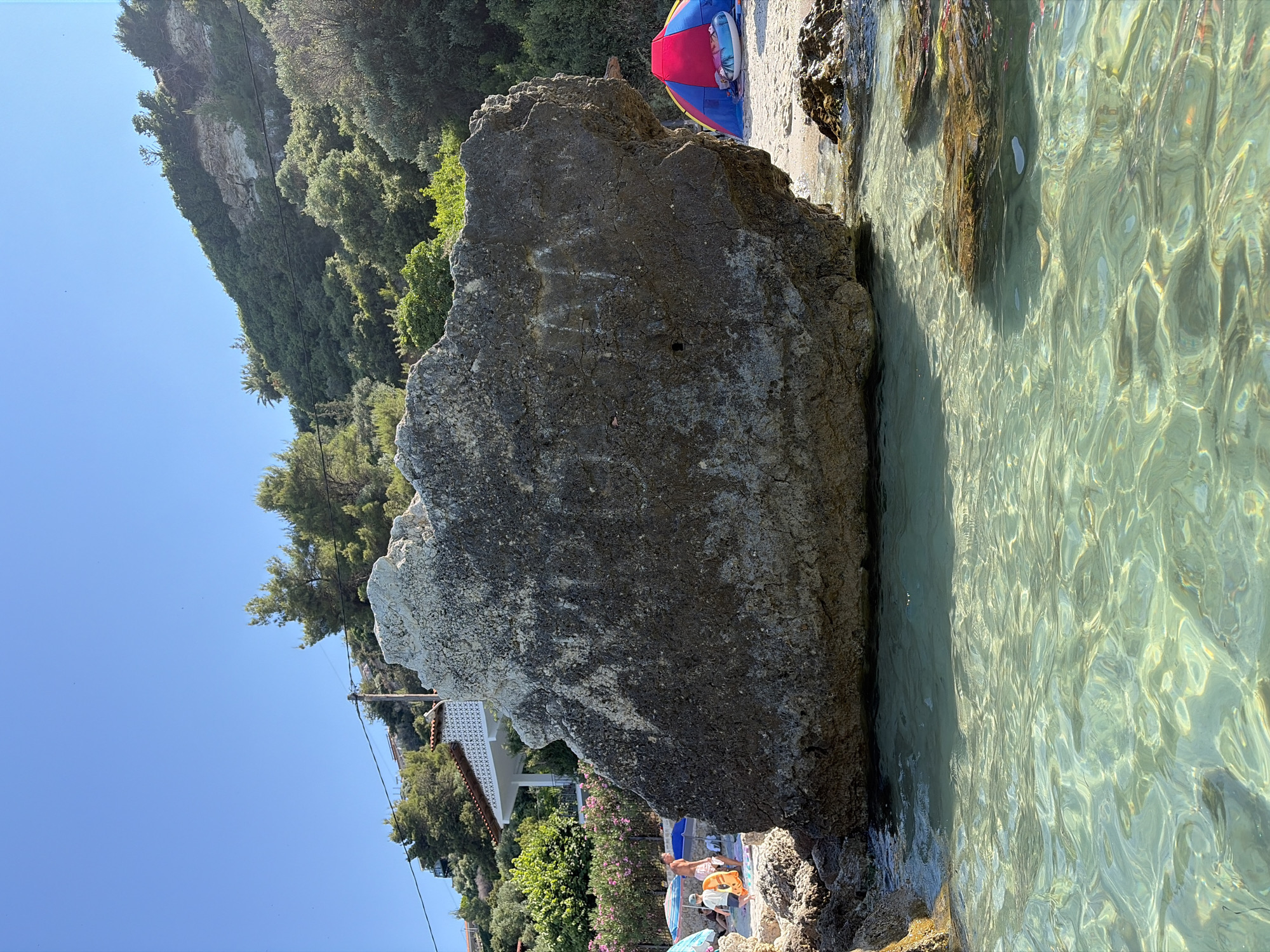 The image depicts a beach scene with a large rock in the foreground partially submerged in clear, shallow water. The water is illuminated by sunlight, creating a shimmering effect on the sandy bottom. Behind the rock, several people are visible on the beach, some relaxing on towels and loungers, while others are near the water's edge. A colorful beach umbrella, a child's inflatable toy, and a surf or bodyboard are also present.
The background features a steep, tree-covered cliff face, with lush green foliage and some exposed rock. A white building with patterned tiles and a red-tiled roof is nestled amongst the trees, suggesting a coastal dwelling or resort. The sky is a clear, bright blue, indicating a sunny day, likely late morning or early afternoon due to the quality of the light and shadows.
The overall atmosphere is one of a relaxed summer vacation. The clear water, sunlight, and presence of beachgoers suggest a pleasant and warm environment. No visible text is present in the image. The location is identified as Kassandra Municipal Unit, Greece.