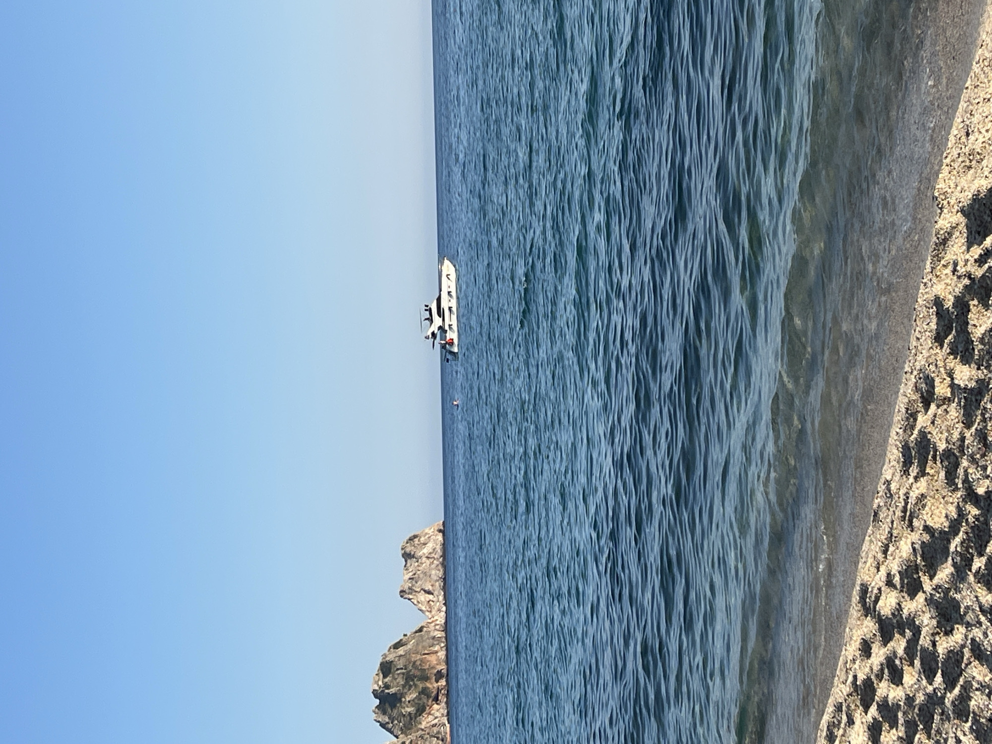 A landscape shot shows the Mediterranean Sea near Alanya, Turkey. The scene is vertically oriented with a clear blue sky on the left. The sea takes up much of the center; textured with gentle waves. A white boat with a few people on board is visible in the sea's center. Near the lower left, a rocky cliff rises from the water. On the right, a sandy beach transitions into the sea. The sand is coarse.