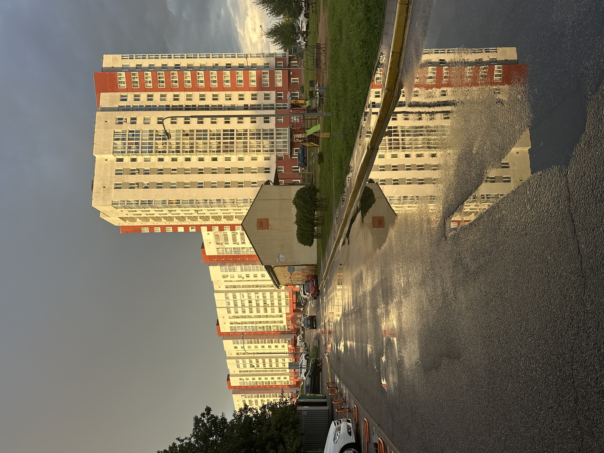 The image shows a street in the city of Minsk, Belarus, with modern apartment buildings on the right side of the frame. The sky is overcast with dramatic clouds, and the setting sun casts a warm, golden light on the buildings. The street is wet, with several large puddles reflecting the buildings and the sky. There are cars parked along the street and in a parking lot to the left of the buildings. In the foreground, there are some orange traffic cones and a white car. To the right of the apartment buildings, there is a small playground with a slide and swings. The overall scene is one of urban living, with a hint of impending rain and the beauty of a sunset.