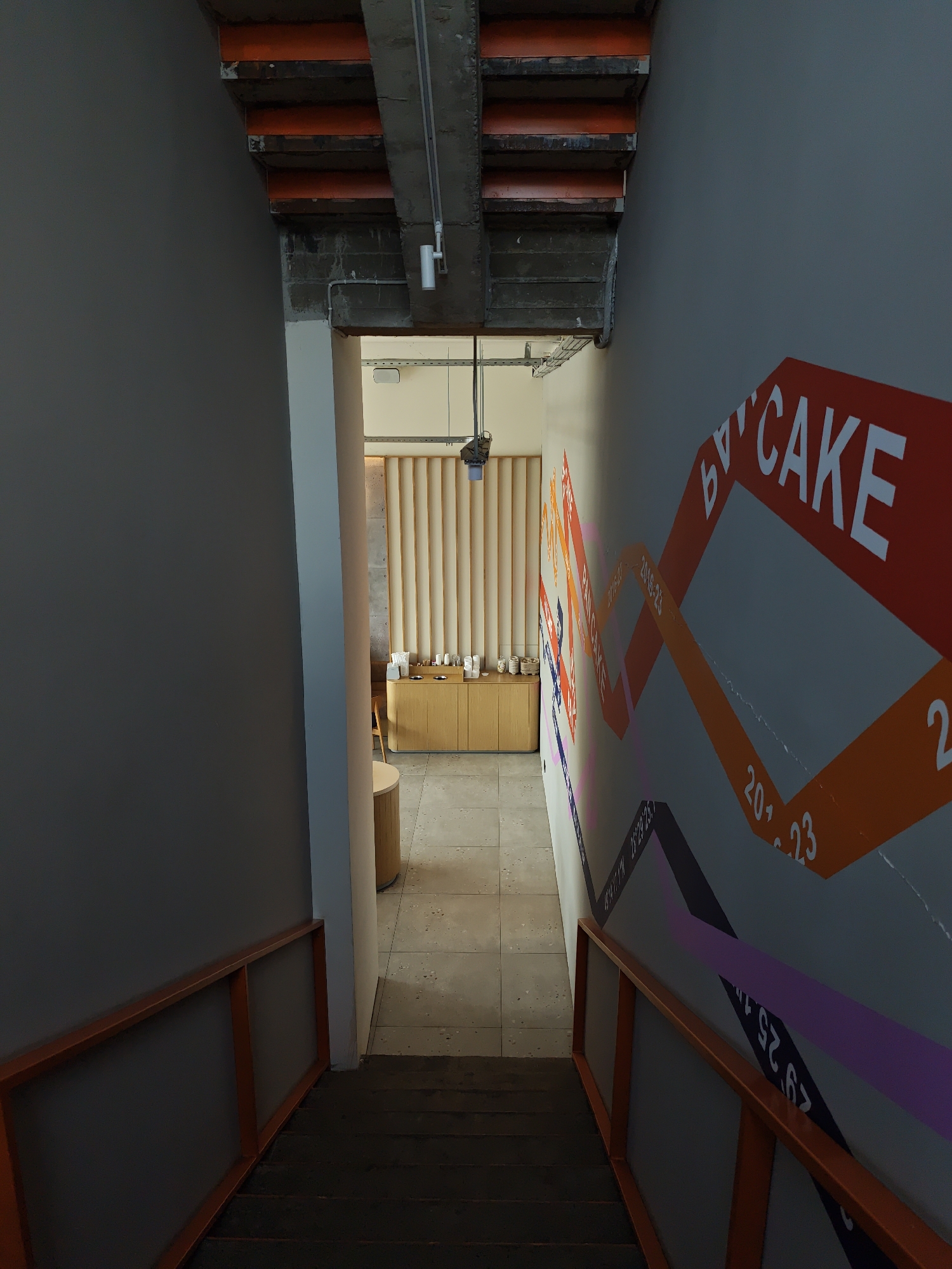 The image depicts the interior of a building, likely a commercial space, in Vinnytsia, Ukraine. The view is down a set of stairs with orange railings. The walls are grey. The wall to the right has large graphic signage displaying the word "CAKE" in a stylized manner, along with other text and graphical elements. The bottom of the staircase leads to an open area with a counter or display case set against a beige, vertically-paneled wall. Various items are displayed on the counter. A cylindrical structure is visible to the left of the counter. The ceiling is a combination of concrete and exposed infrastructure. The flooring appears to be light grey tile. The scene is well-lit, suggesting an indoor setting during daylight hours. There are no people visible in the frame.