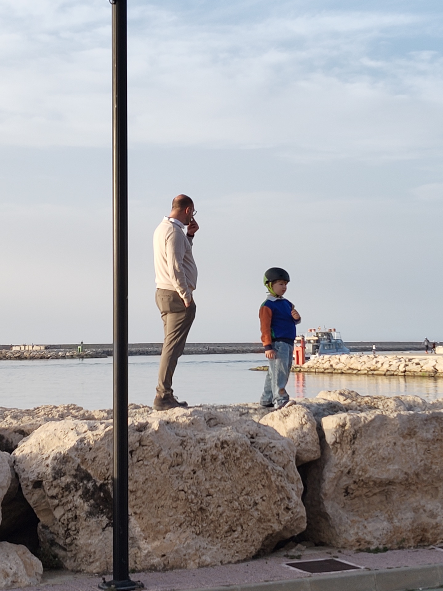 A photo shows a man and a child standing on large rocks at the edge of the sea. The man is on the left side of the frame, wearing a light-colored sweater, brown pants, and glasses; he is holding something to his face and appears to be talking. To his right stands a child wearing a blue shirt with brown sleeves, jeans, and a helmet. In the background is water, a pier, and some buildings. A tall, black pole stands near the man. This scene is from Dénia, Spain, where stone breakwaters and a marina are visible along the coastline.