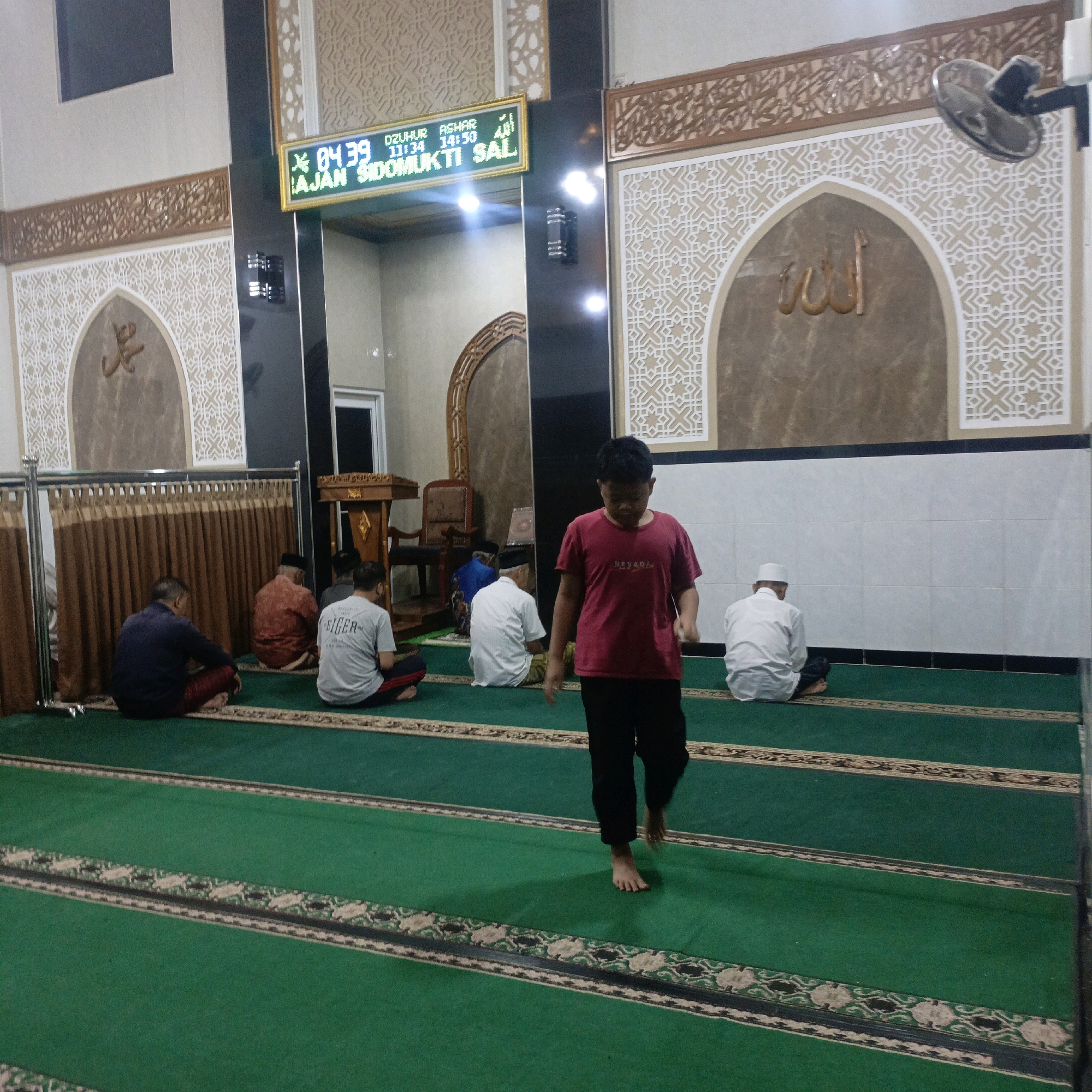 The image depicts the interior of a mosque, likely located in Salatiga, Indonesia, as indicated by the text on the digital display. The scene captures individuals engaged in prayer on a green carpeted floor, which is marked with prayer lines. The walls feature Islamic decorative patterns and calligraphy, with a prominent arched niche containing Arabic script. A digital display showing time and prayer schedules is visible on the wall. A young boy in a maroon t-shirt and black pants is walking across the prayer area. Several men are seated on the floor in prayer positions, facing the qibla. The environment appears to be dimly lit, with the primary light source coming from the digital display and some ambient ceiling lights. A ceiling fan is also visible in the upper right corner. The overall atmosphere suggests a place of worship during prayer time.