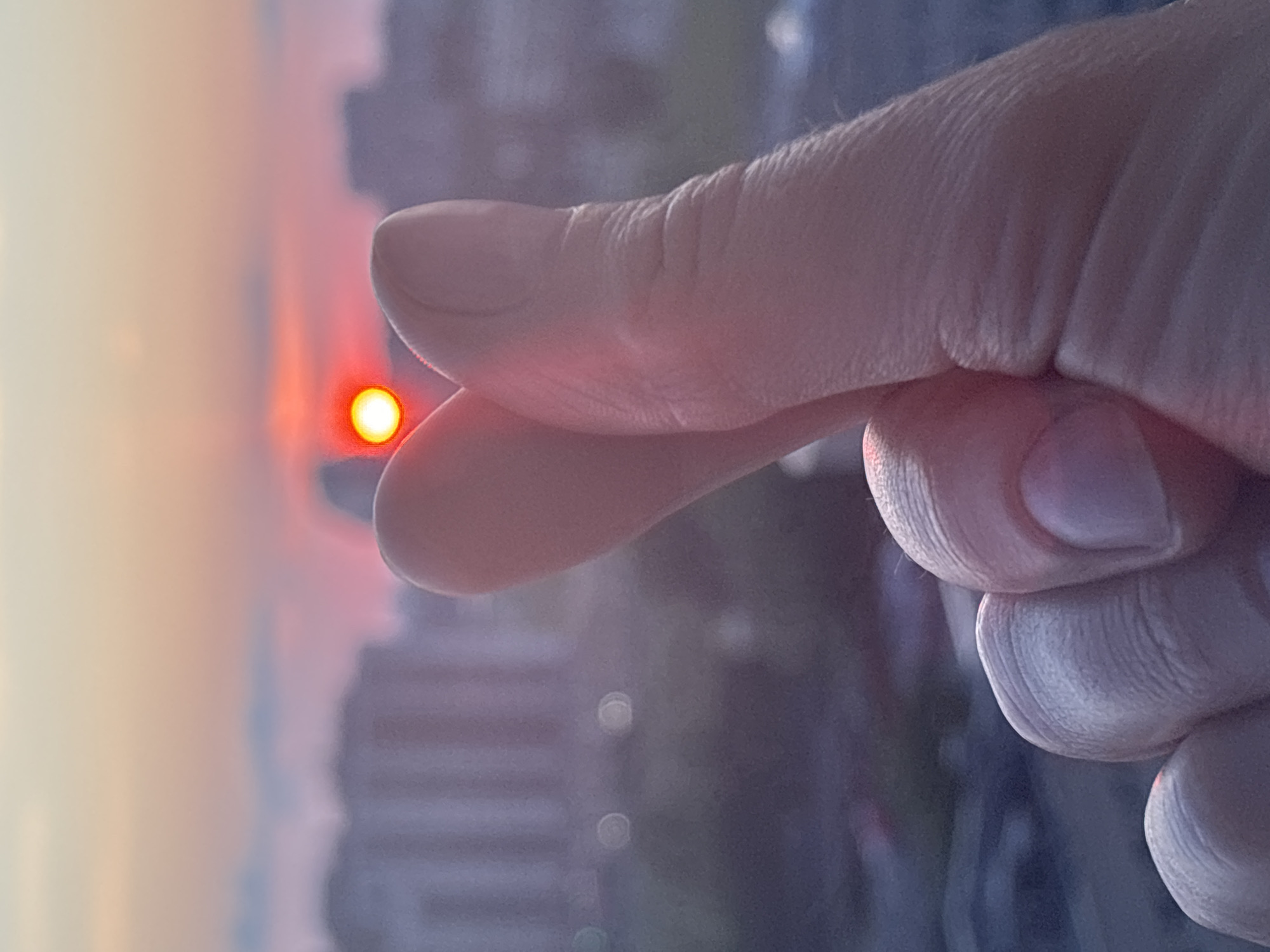 A close-up, shallow depth-of-field shot features a person's hand in the foreground, forming a heart shape with their thumb and index finger. The sunset, a vivid red orb surrounded by a halo of pink and orange light, is visible behind the hand. The background is a blurred cityscape with buildings silhouetted against the colorful sky, hinting at a location in Unknown, Russia, likely from an elevated vantage point.