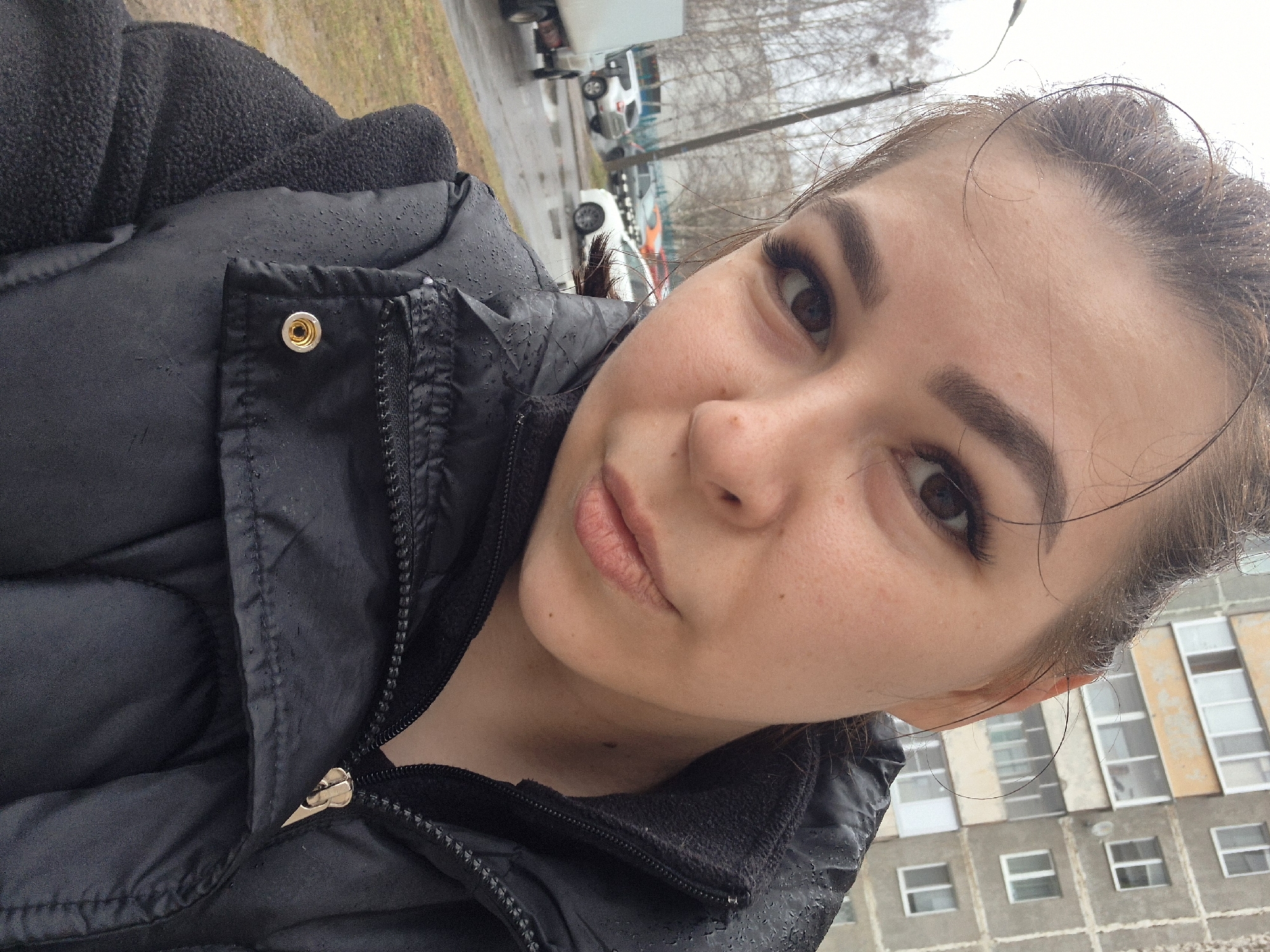 A close-up selfie depicts a woman with brown eyes and hair, wearing a black puffer jacket. The image appears to have been taken outdoors on a wet day, with visible water droplets on her hair and clothing. In the blurred background, there are cars, trees, and a building with multiple windows. Based on these details, the location is likely in the city of Tobolsk, Russia. The interaction is primarily with the camera, as the woman is posing for the selfie. The environment is overcast and damp.