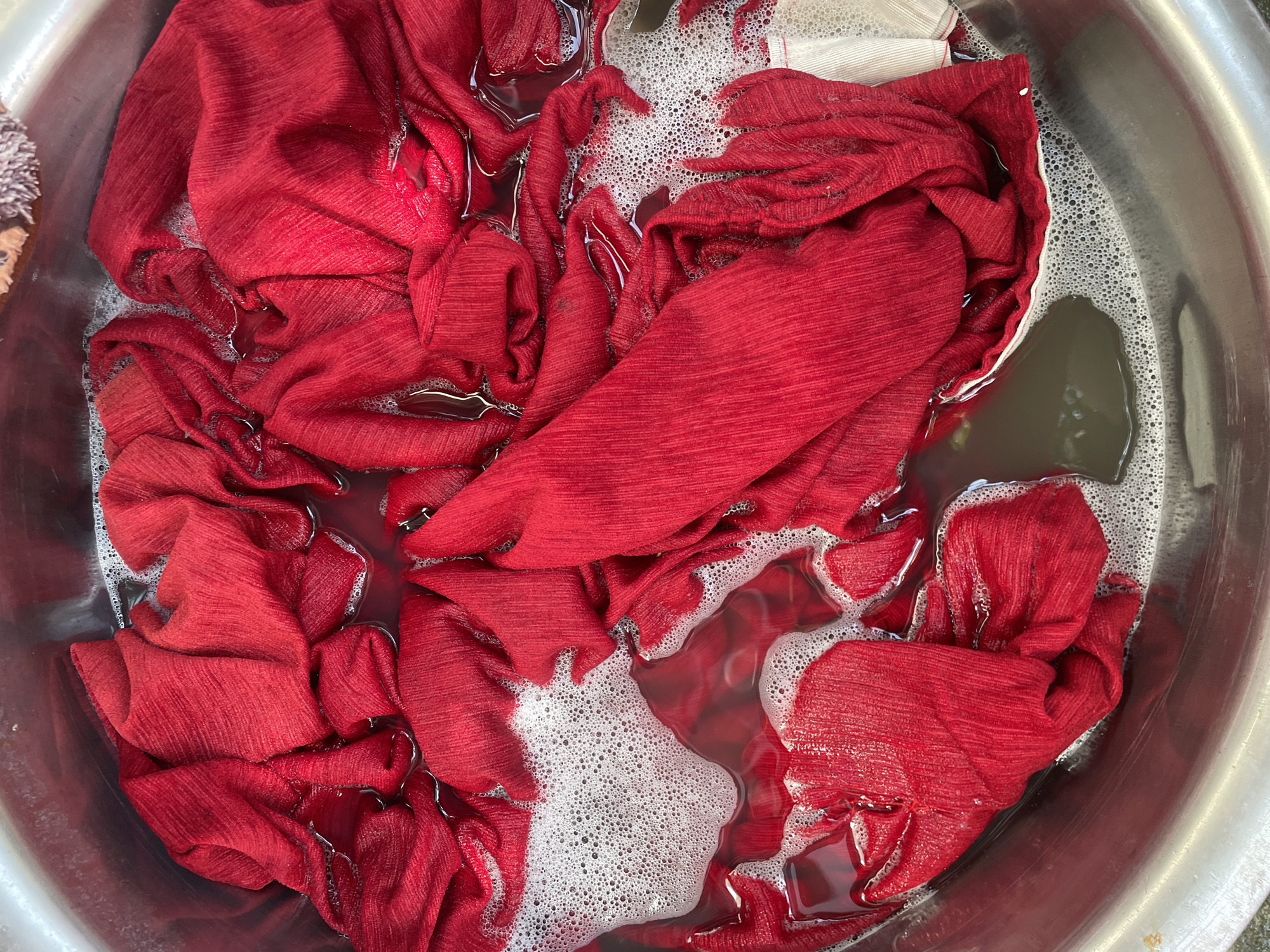 The image presents an overhead view of vibrant red fabric soaking in a metal basin, likely during a hand-washing process. The scene is dominated by the crumpled and folded red textile, which exhibits a subtle crinkled or ribbed texture. Several pieces of this red fabric are submerged in murky water, with white soap suds and bubbles visible on the surface, particularly concentrated in the upper right and central areas of the basin.
The water itself appears discolored, showing a reddish tint, possibly from dye bleeding from the fabric, combined with a darker, brownish hue, indicating the presence of dirt or impurities being washed out. A small portion of a white fabric item can be seen in the upper-right corner, partially covered by the red cloth and foam. The metal basin has a polished, reflective rim and bottom, suggesting it is made of stainless steel or a similar material.
The activity depicted is the soaking or washing of clothing, a common domestic chore. The presence of soap and foamy water confirms this. No people are visible in the frame. There are no discernible location cues within the image itself, though the activity could be taking place in a household setting in Davao City, Philippines, as per the provided context. No text is visible in the image.