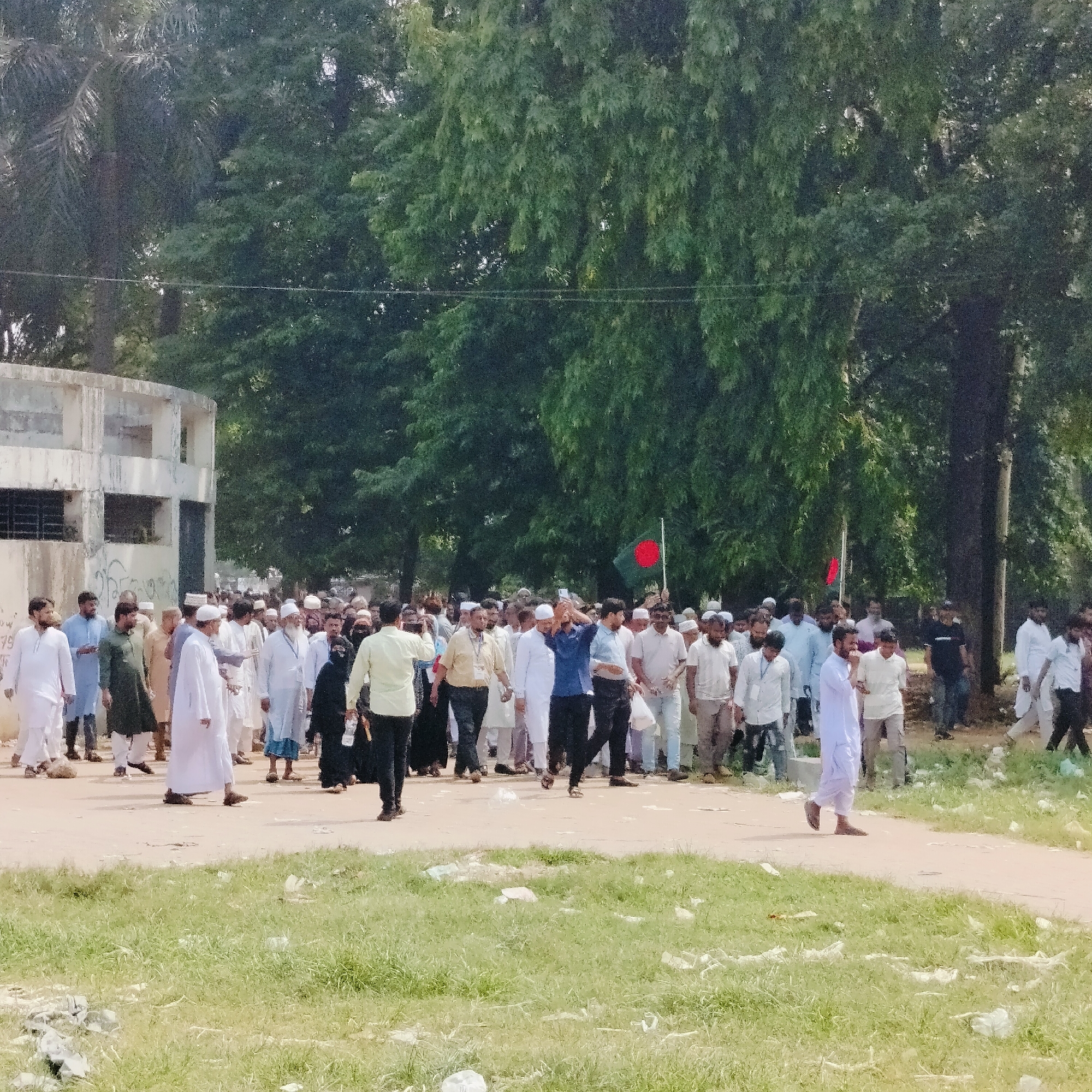 A large crowd of people, primarily men, are walking along a road in Dhaka, Bangladesh. Most of the men are wearing traditional white Muslim attire, with some in more casual clothing. A few individuals are carrying Bangladeshi flags. The road is unpaved and littered with debris. Buildings and trees are visible in the background.