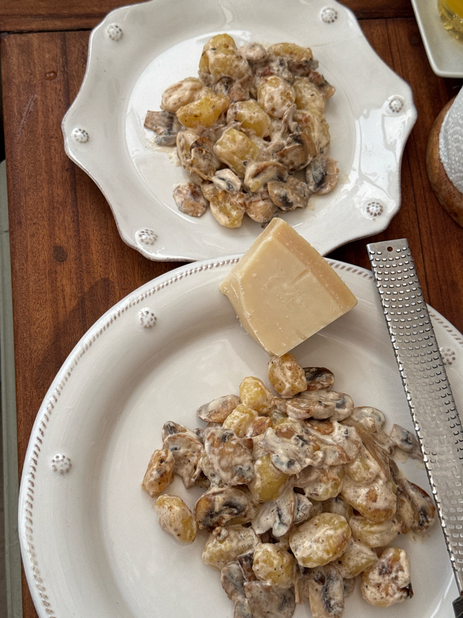 Two white plates of gnocchi with mushrooms in a creamy sauce are arranged on a wooden table. A wedge of parmesan cheese and a grater are placed on the bottom plate. The top plate is slightly out of focus. The lighting suggests an indoor setting, possibly a home or restaurant. The specific location in Essaouira, Morocco, cannot be determined from the image.