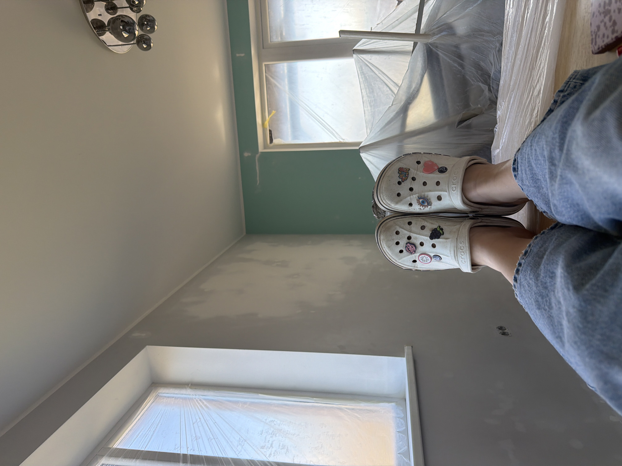 A first-person perspective shows a pair of legs clad in denim jeans, with white Crocs on the feet, standing inside a room in what appears to be a building under renovation. The room has walls that are partially painted in shades of grey and green, with exposed patches. There are two windows, covered with transparent plastic sheets. Building materials are partially covered with plastic sheeting. A light fixture is visible on the ceiling. The location information is not discernible in the image.