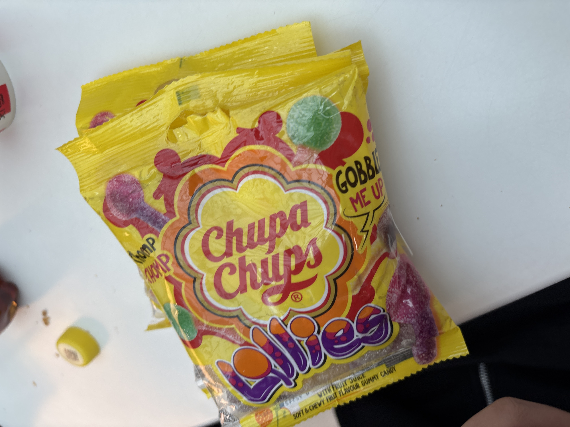 A close-up shot of a bag of "Chupa Chups Lollies" gummy candy, set against a white surface with portions of other objects in the periphery. The bag is yellow with a clear section revealing the colorful gummy candies inside. The "Chupa Chups" logo is prominently displayed. The candy is labeled as "SOFT & CHEWY FRUT FLAVOUR GUMMY CANDY" and "WITH FRUIT JUICE". The phrase "GOBBLE ME UP" is also visible on the packaging. A yellow candy piece is visible on the left side.