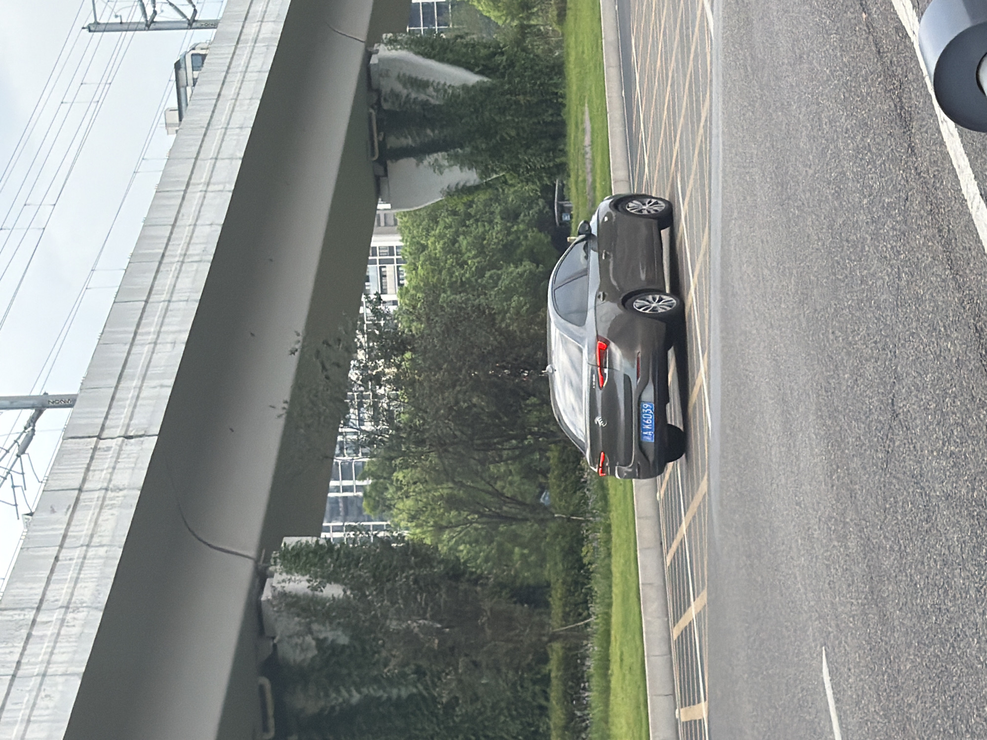 In the city of Jiangjin, China, a gray sedan with a license plate reading "AK6039" is parked on a paved road under a large concrete overpass. The overpass has some greenery growing on its supporting pillars. In the background, there are trees and buildings, suggesting an urban or suburban environment. The sky is overcast.