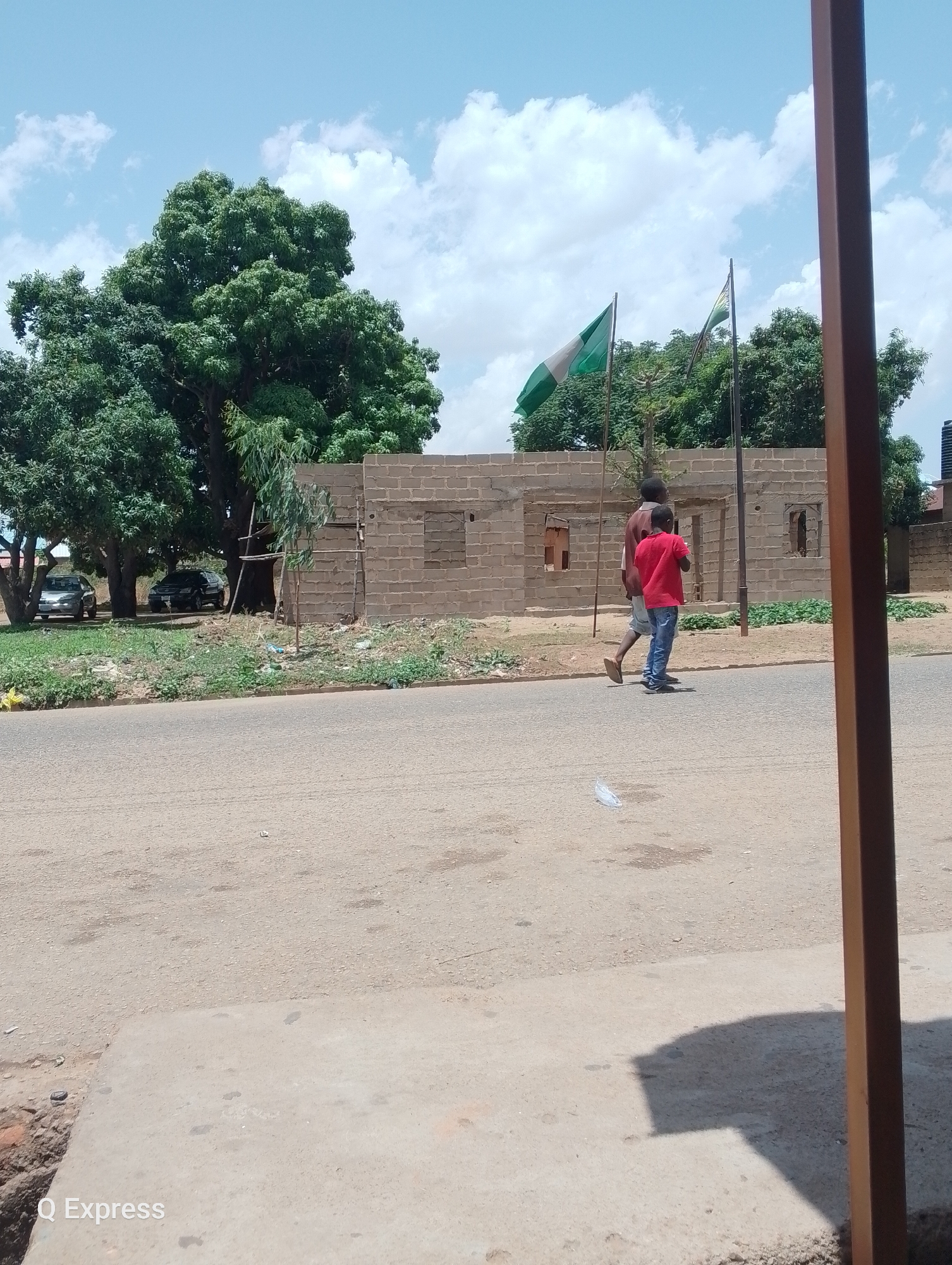 The image shows a street scene in Gura Top, Nigeria, on a sunny day. The foreground is dominated by a paved road, with a partial view of a brown support structure in the right foreground. Two individuals are walking along the road, an adult male and a younger boy wearing a red shirt. Behind them, a building under construction with visible window openings and two Nigerian flags flying nearby is visible. A cluster of large trees is to the left of the building. Two cars are parked near the trees. The sky is blue with scattered white clouds. A water tank is on top of a building to the right. "Q Express" is printed in the lower left corner.
