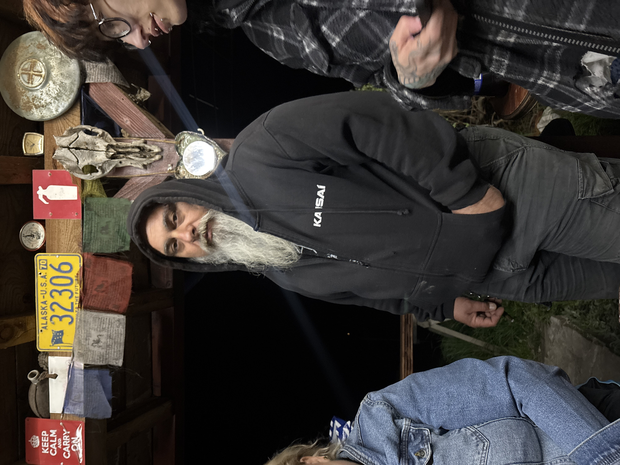 The image shows a scene that appears to be indoors, potentially in a rustic or eclectic setting. There are at least three people visible, including a man with a long white beard wearing a black hoodie with the word "KAISAI" printed on it. He is positioned in the center of the image, looking towards the camera. Above him and to the left, there are several items attached to a wooden structure, including what looks like a license plate from Alaska with the number "32306", a possible animal skull, a clock, and some decorative items or flags. To the right of the man, there is another person wearing a plaid shirt, with part of their arm and hand visible. Below the man, there is a person wearing a denim jacket. The background is dark, and there is a beam of light shining upwards. There are no specific details in the image to confirm if this is in the city of Verinsko, Bulgaria.