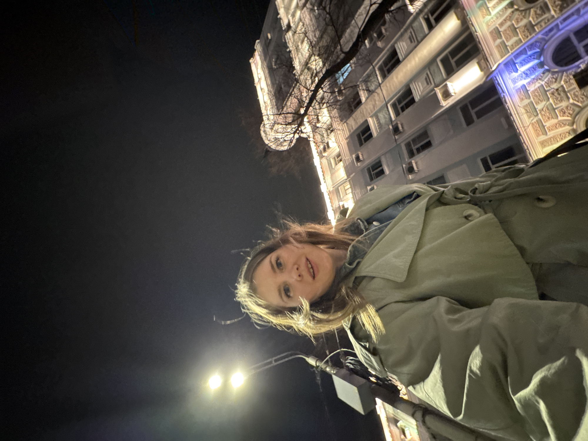 A woman with fair skin and blonde hair is photographed at night, facing the camera with a slight smile. She wears a light green trench coat over a denim jacket. The photograph's perspective is from below, looking upwards. A tall, illuminated building with many windows and ornate architectural details forms the backdrop behind the woman. Bare tree branches are visible in front of the building. A street lamp illuminates the scene in the lower part of the frame.
The location is in Perm, Russia, near the Permyak Hotel.