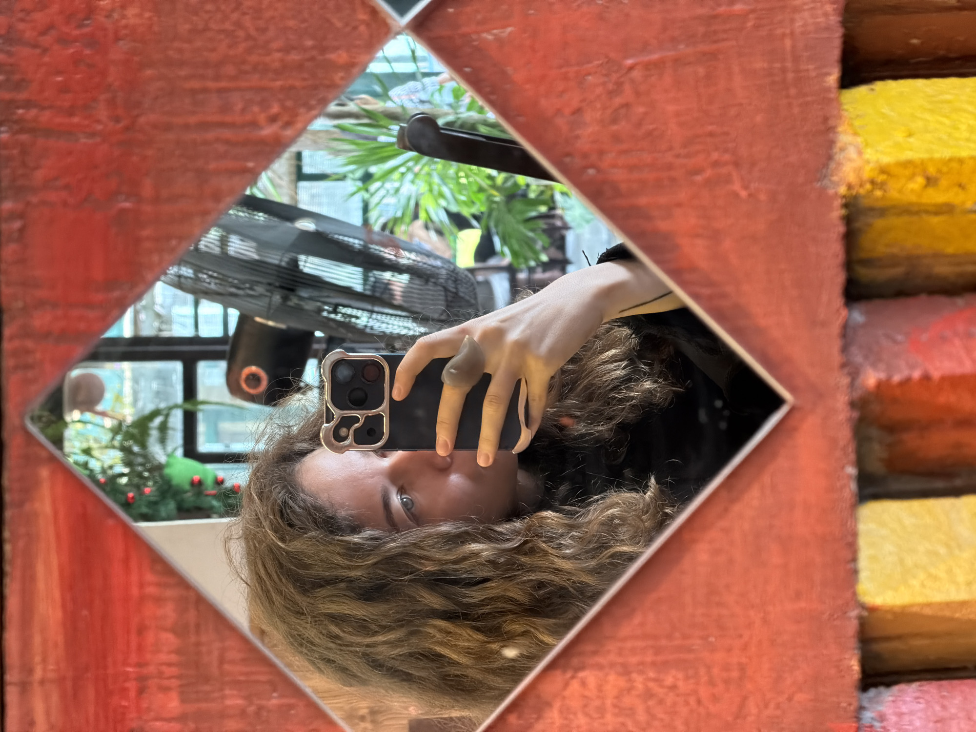 A mirror selfie is being captured by a person in a diamond-shaped mirror, set within a textured, reddish-orange frame. The individual, seen partially in reflection, has curly light brown hair and one visible eye, which appears to be looking towards the camera. Their hand holds a smartphone with a distinctive camera module, pointed towards the mirror.
In the reflections visible through and around the person, several elements of the background are discernible. A black standing fan is seen in the upper left of the mirror, alongside lush green foliage and what appears to be a small potted plant resembling a Christmas tree adorned with red berries. Windows or glass panels are also reflected, suggesting an indoor setting with natural light, indicative of daytime.
Outside the mirror's immediate border, on the right side of the red frame, a stack of rectangular objects is visible, painted in vibrant blocks of yellow, orange, and a lighter pink or red hue. The textured, painted surfaces suggest an eclectic or decorated interior space, possibly a cafe or a uniquely styled room in Đà Nẵng, Vietnam. No visible text is present, and the person's expression is neutral, focused on taking the photograph.