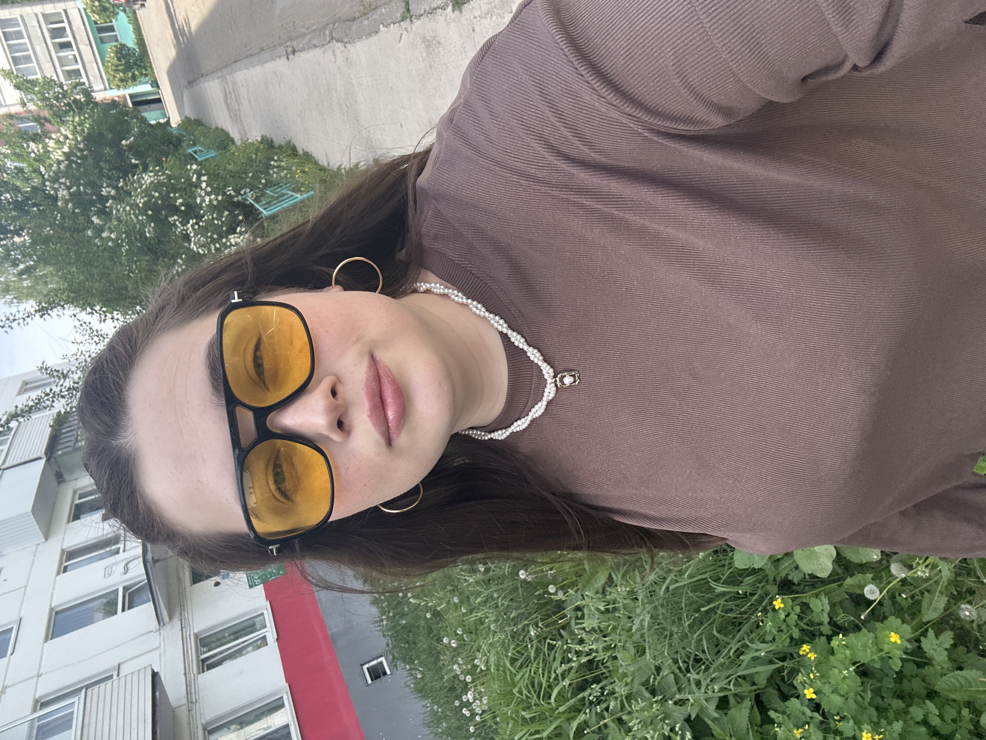A young woman is standing outside in what appears to be a city setting, likely in Chaikovsky, Russia. She is wearing yellow-tinted sunglasses, hoop earrings, and a pearl necklace, and a brown, long-sleeved shirt. A building, possibly an apartment block, is visible in the background, featuring white walls, windows, and red and gray accents. The ground is covered with greenery, including grass, wildflowers, and dandelions. A tree or shrub is also visible on the left side of the image. The time of day is likely daytime due to the visible sunlight.