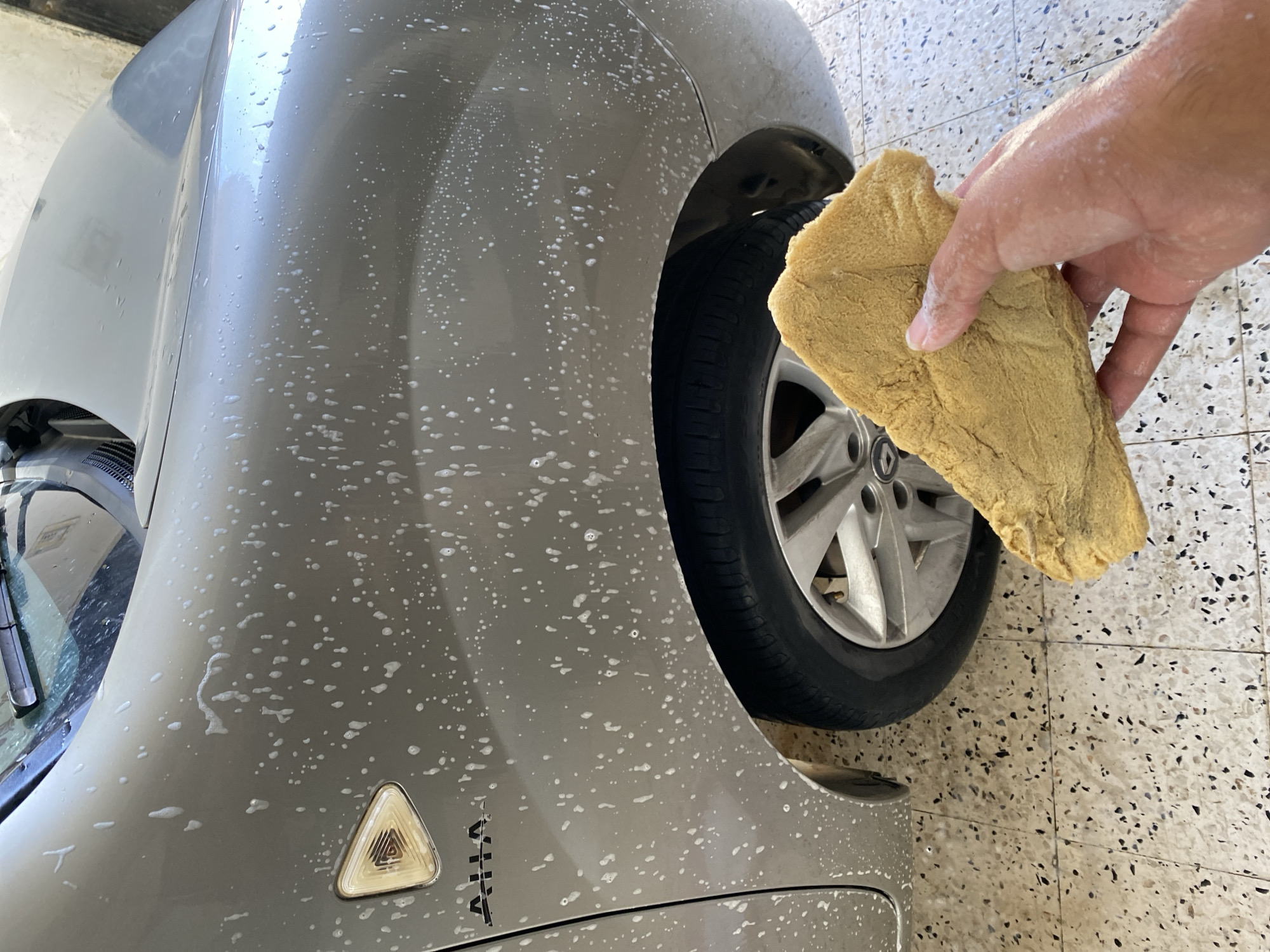 The image shows a close-up of a car being washed, with a sponge held by a hand in the foreground. The car is a light gray color and is covered in soap suds. The sponge is yellow and appears to be made of a porous material. The scene is set on a tiled floor, suggesting an outdoor area, likely a driveway or a car wash bay. The overall impression is of a routine car cleaning in progress. The location is identified as Baraki, Algeria.