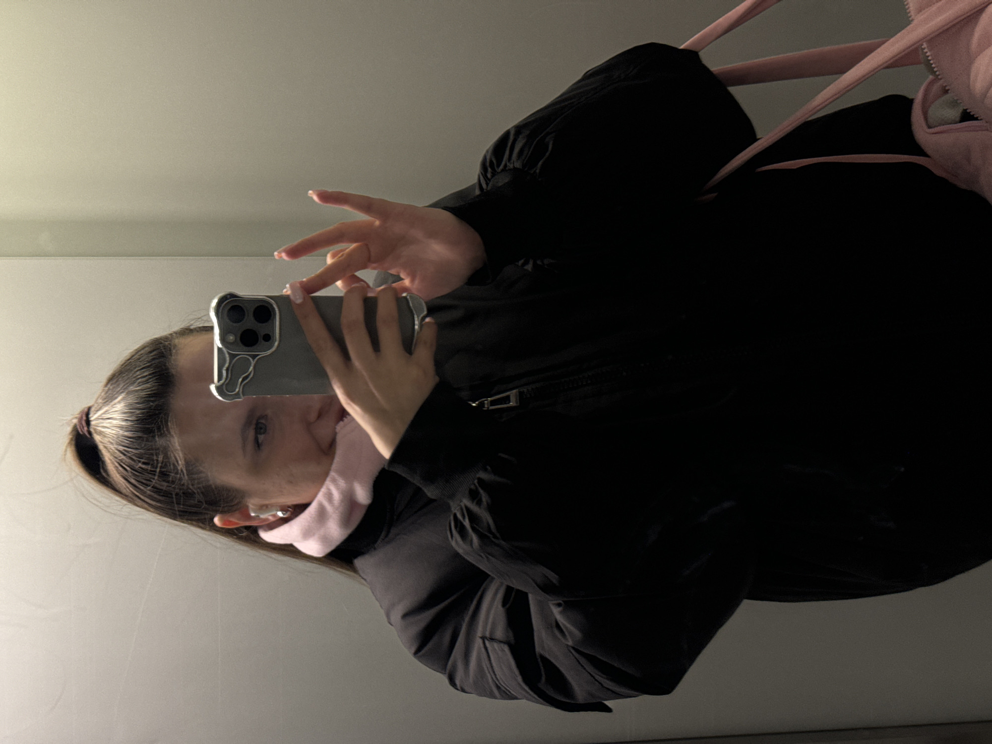 A woman is taking a selfie in what appears to be an elevator or changing room. She is wearing a black jacket and a light pink scarf or neck warmer. Her hair is pulled back in a ponytail. She is holding a cell phone up in her right hand to take the photo. A pink bag is hanging in the background. The walls are a light grey or white.
I am unable to provide the specific location in Odintsovo, Russia.