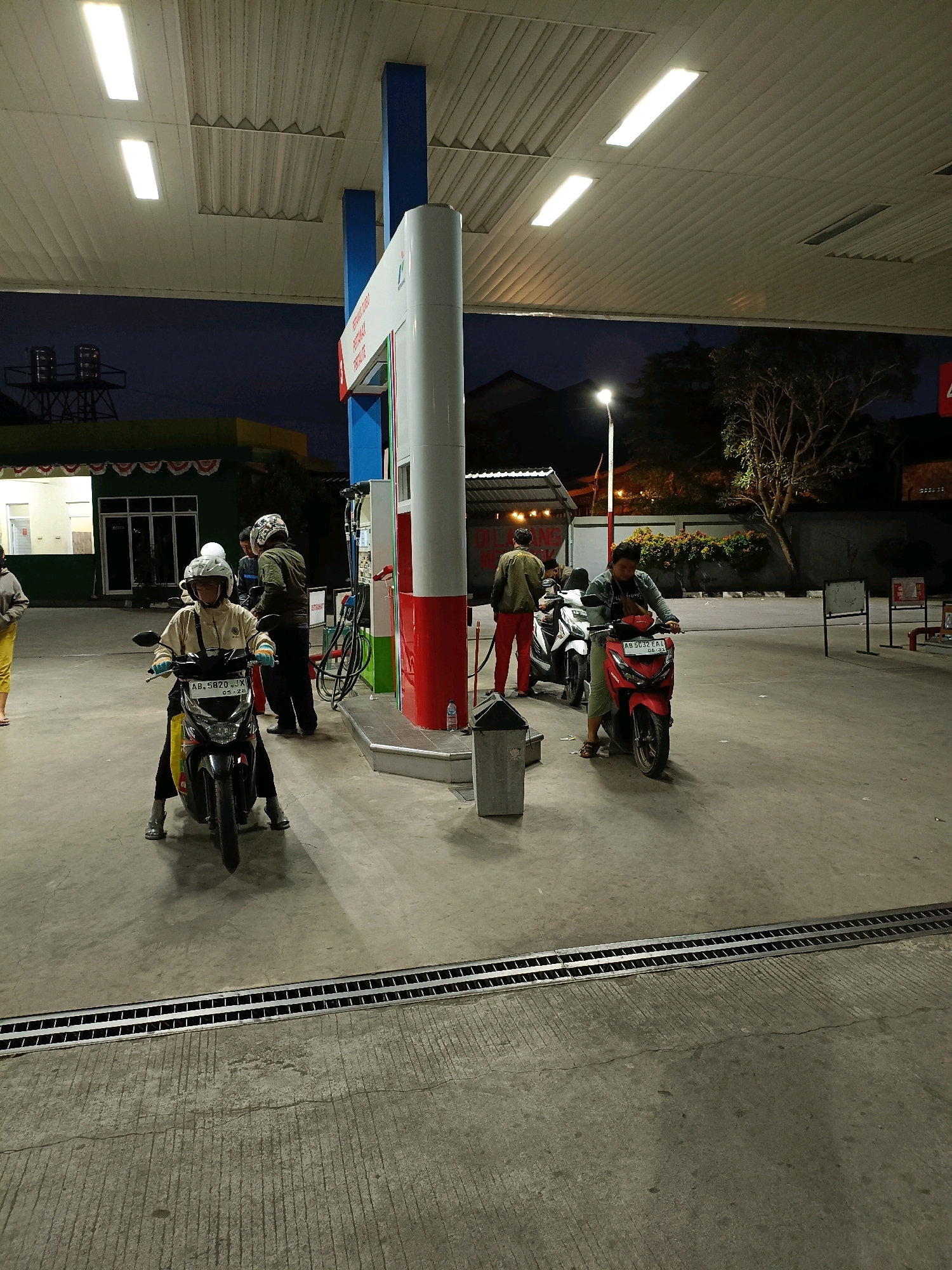 At a gas station in Maguwoharjo, Indonesia, at dusk, several people are seen with their motorcycles. The scene is illuminated by overhead fluorescent lights and a streetlamp in the background. The ground is paved concrete with a metal drainage grate running across it. A red and white gas pump stands prominently in the center of the frame. In the foreground, a person wearing a yellow jacket and white helmet is seated on a black motorcycle, its license plate reading "AB 5820 JX". To the right, another person on a red motorcycle, license plate "AB 5032 EA", is also visible. Further back, other individuals and motorcycles are present near the gas station buildings and canopy. The sky is a deep blue, indicating the transition from day to night.