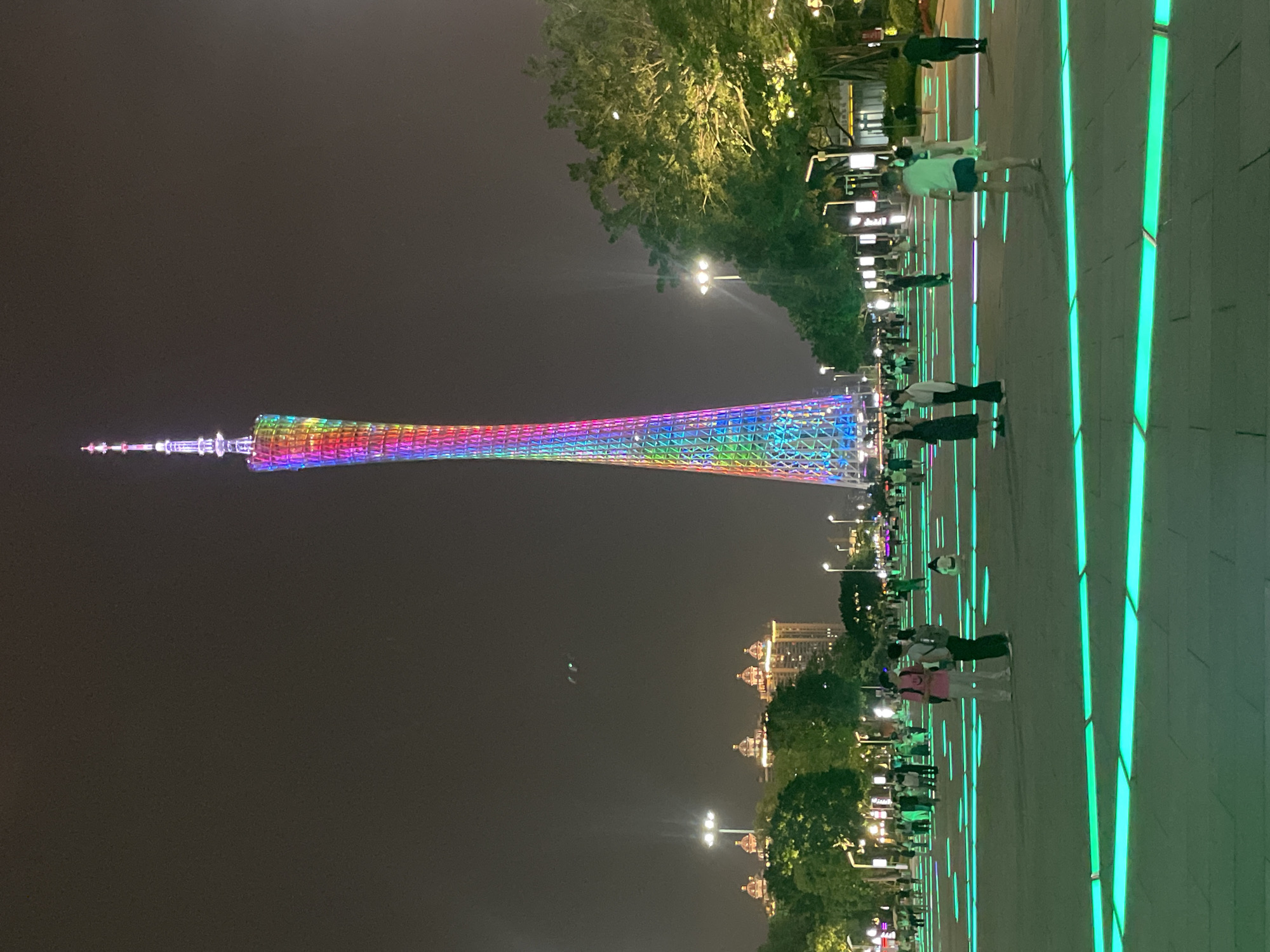 The image depicts the Canton Tower in Guangzhou, located in the Tianhe District, China, illuminated with a colorful array of lights at night. A wide, paved walkway with embedded green and other colored lights runs along the right side of the image, where numerous people are walking and standing. Street lights and buildings can be seen in the distance.