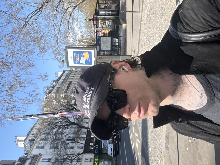 A single male subject is depicted in a medium close-up, facing left, in an urban outdoor environment under clear blue skies. The subject wears a distressed black baseball cap with "JUST IS WHERE ARE STANDING" embroidered in white, large black rectangular sunglasses, and a silver earbud in his right ear. Visible tattoos cover his neck, including the number "1711," and extend onto his upper chest. He wears a black t-shirt and a dark, possibly suede, jacket with a black strap from a bag over his right shoulder. A silver necklace with a small pendant is visible.
The background conclusively identifies the location as Paris, France, featuring a distinct "METRO" station entrance with its characteristic sign and glass structure to the right. To the left of the Métro entrance, a public information panel displays "PARIS" at its top. Classic Parisian Haussmannian buildings with light facades and numerous windows line the street. A "PHARMACIE" sign is visible on a building in the far left background. A street with a white car and a scooter is present. Bare tree branches, devoid of leaves, extend across the upper portion of the frame. The ground is paved with urban cobblestones or similar material.