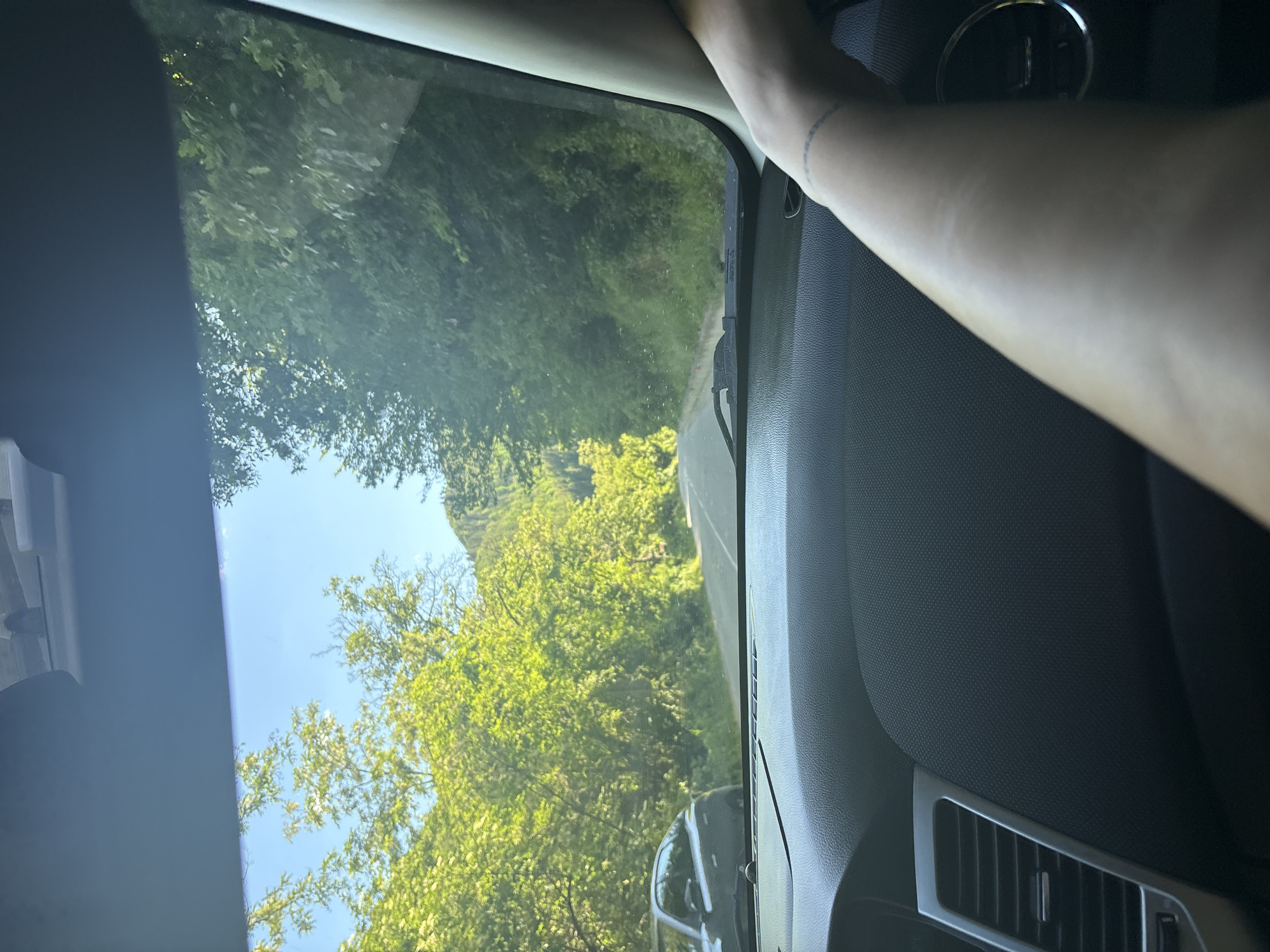 The image is taken from inside a car looking out through the windshield. The scene depicts a winding road passing through a dense forest. The sun is shining brightly, casting dappled light on the lush green foliage. On the left side of the road, a dark-colored car is parked. The dashboard of the car is visible in the foreground, along with a person's arm and hand resting on the steering wheel. The arm has a subtle tattoo around the wrist. The overall impression is that of a scenic drive on a sunny day in a rural area. The location is Hvoina, Bulgaria.