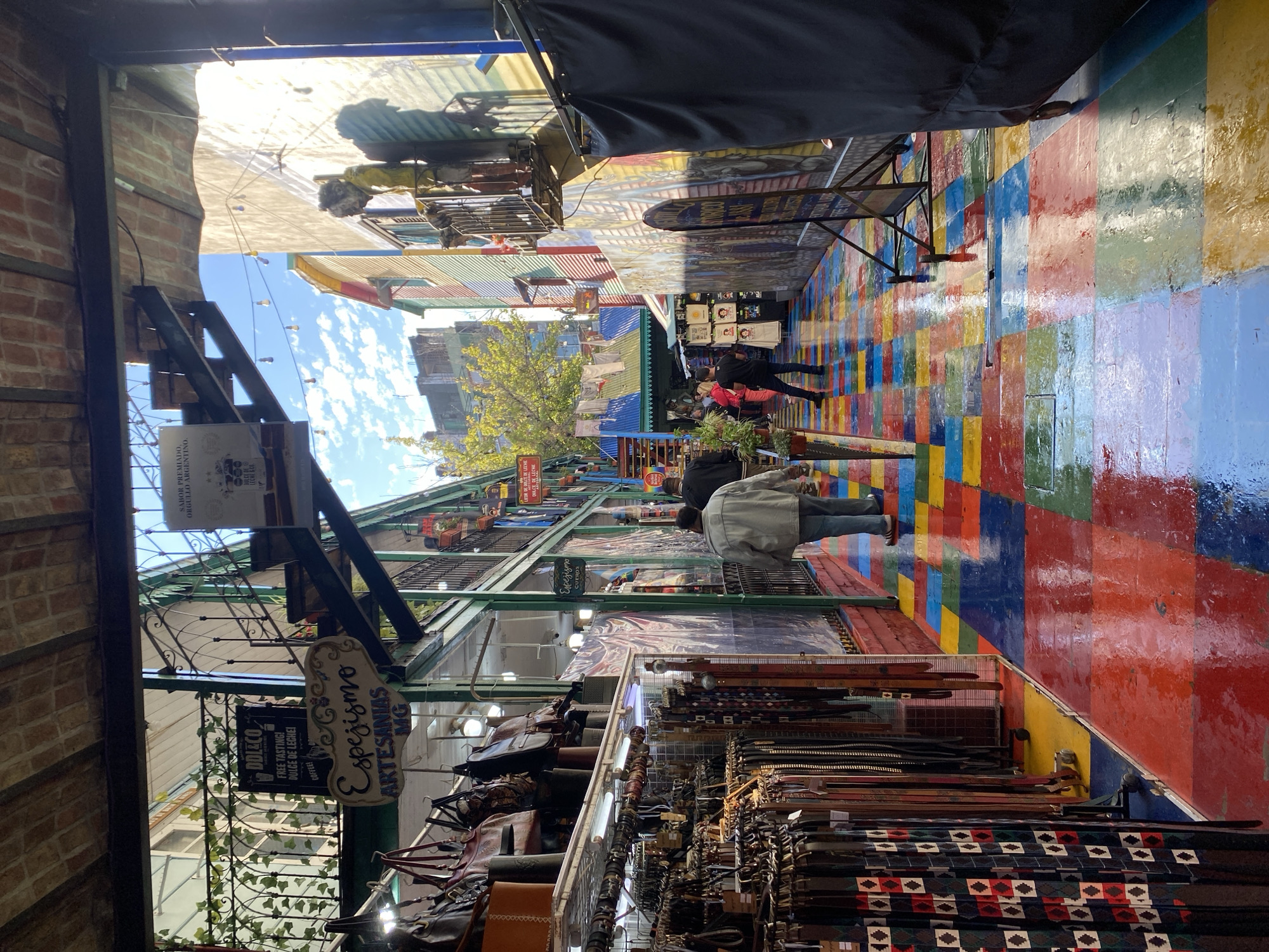 This vibrant image captures a bustling, colorful outdoor market or shopping alley, likely in Caminito, La Boca, Buenos Aires, Argentina. The ground is a striking feature, paved with large, glossy squares in a mosaic of bright red, yellow, blue, and green, appearing wet and highly reflective, possibly from recent rain or cleaning. Above, patches of blue sky dotted with white clouds indicate it is daytime.
Several people are seen casually strolling through the alley, which is lined with shops and market stalls. Two individuals are prominent in the middle ground, one in a dark jacket and another in a light-colored jacket, both wearing jeans. On the right side of the path, merchandise is displayed, featuring numerous belts with diverse patterns and buckles, alongside what appear to be leather bags and other accessories. The left side showcases other stalls, some with hanging items and signs.
The architectural style features rustic elements like exposed brick walls and corrugated metal roofs. String lights with bare bulbs hang overhead, suggesting the area might be lit for evening events or decorative purposes. Greenery, possibly vines, grows on metal grilles, adding a touch of nature.
Visible text on various signs includes: "ODLECO FREE TASTING! DULCE DE LECHE! COFFEE!", "Espejismo ARTESANIAS MG", "Espejismo CUEROS", and "SABOR PREMIADO, ORGULLO ARGENTINO." These signs suggest local artisan crafts and culinary delights like dulce de leche. The scene conveys a lively, colorful, and inviting atmosphere typical of a popular tourist destination.