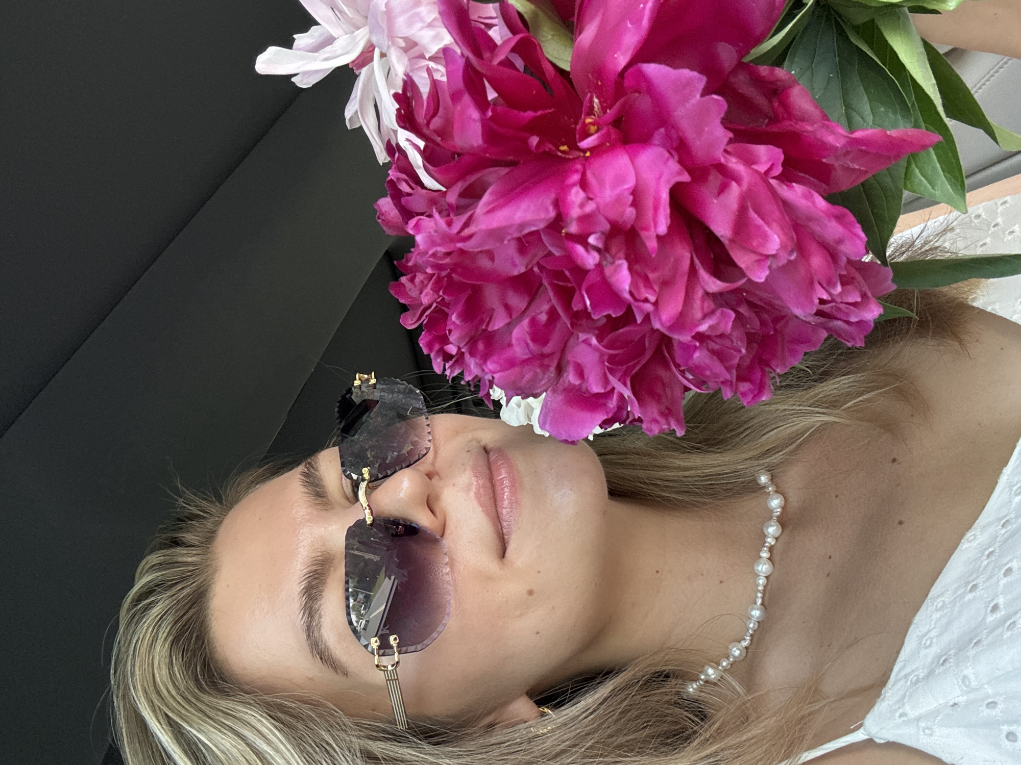 A young woman with blonde hair and sunglasses is holding a bouquet of bright pink and white peonies. She is wearing a white eyelet top and a pearl necklace. The background is dark and appears to be the interior of a car. The woman is smiling slightly and looking towards the flowers. The image is taken from a close-up perspective, focusing on the woman's face and the flowers. The lighting is natural and appears to be daylight. The overall impression is one of natural beauty and a moment of enjoyment. The location is Unknown, Unknown.