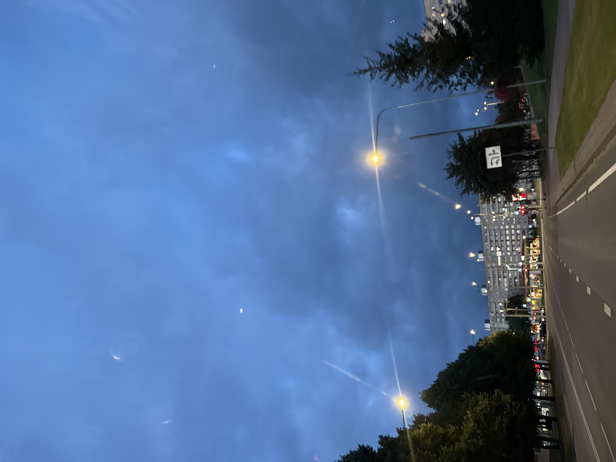 The image captures a street scene in Tallinn, Estonia, likely during dusk or early evening, given the illumination of streetlights and the darkening sky. The perspective is from a moving vehicle on a multi-lane road. On the left, a line of trees borders the road, with a hint of residential buildings or shops behind them. The road itself has clear lane markings. To the right, a sidewalk area is visible, with grass and some trees, including a prominent evergreen tree. In the background, several tall apartment buildings are visible, characteristic of Soviet-era architecture, and a gas station with illuminated pumps adds to the urban landscape. The sky is a deep blue with visible clouds and what appear to be contrails from an aircraft, as well as a couple of bright, small points of light that could be stars or distant lights. A traffic sign showing directional arrows for different lanes is also present on the right side of the road.