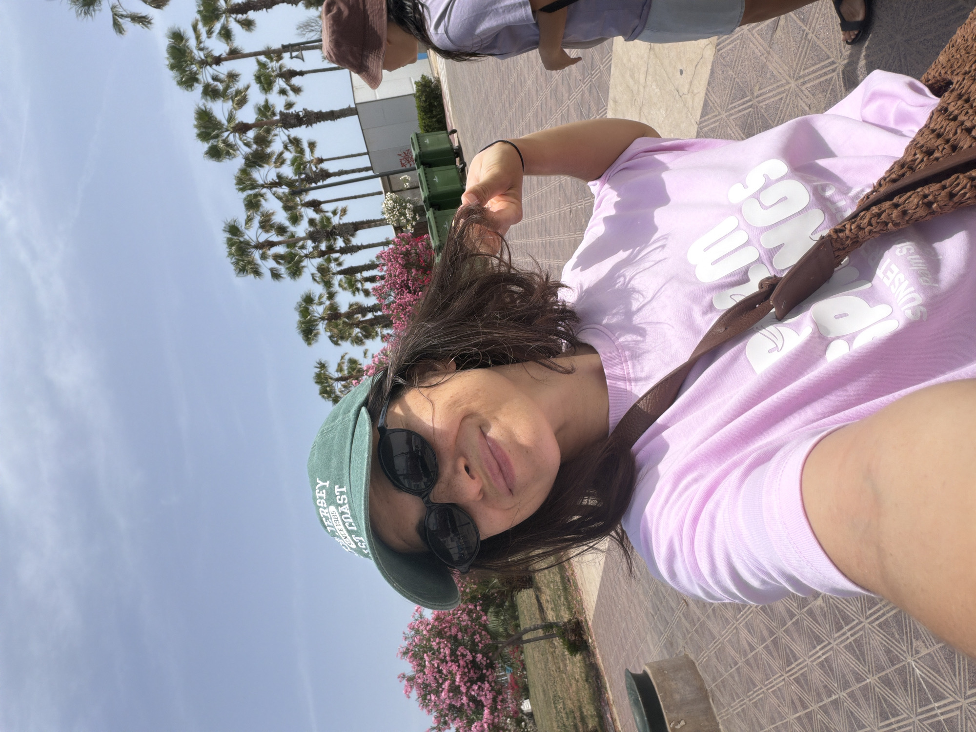 A selfie is taken of a woman wearing a green baseball cap and sunglasses, with a pink t-shirt and a woven brown bag over her shoulder. She is smiling and touching her hair. In the background, palm trees line a street with green garbage bins and a building. Another person wearing a brown bucket hat and a grey t-shirt is visible on the right. The ground is paved with patterned tiles, and there are pink flowers in the background.