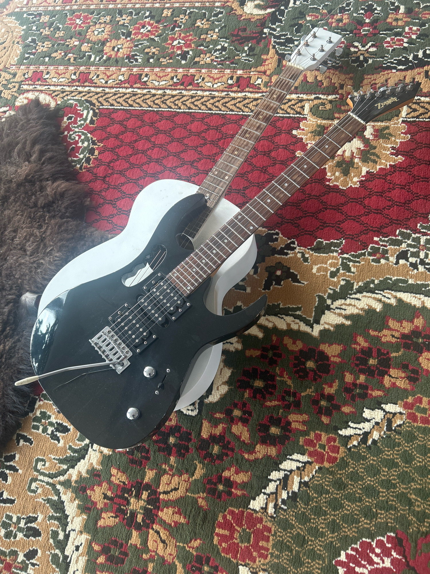 Two electric guitars, one black and one white, are laid diagonally across a richly patterned rug. The black guitar is in the foreground, featuring six strings, two pickups, and a tremolo bar. The white guitar is partially visible underneath, with its headstock and neck extending upwards. A dark, shaggy material, possibly fur, is positioned to the left of the guitars. The rug displays intricate floral and geometric designs in colors of red, green, brown, and cream. The scene is indoors, with no discernible environmental context that would suggest a specific location in Grozny, Russia.