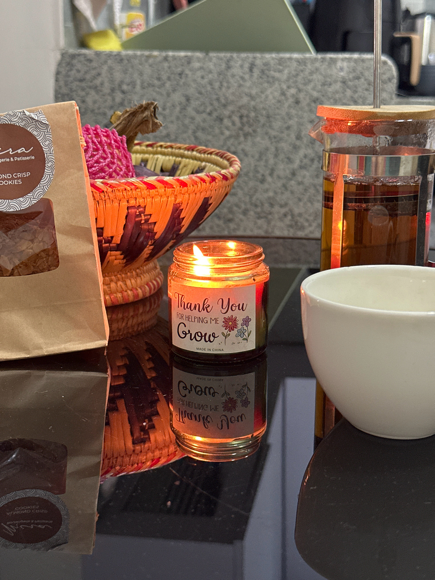 The image depicts a close-up, eye-level shot of a dark, reflective surface, likely a table, on which several objects are placed. In the foreground, a lit candle in a glass jar glows warmly, with a label that reads "Thank You For Helping Me Grow." To the left of the candle, a brown paper bag containing cookies is visible, with a circular label partially obscured. Behind the candle and bag, an orange and purple woven basket holds some items, including a pink knitted object and a dried plant. To the right of the candle, a glass teapot filled with amber liquid, possibly tea, stands next to a white ceramic bowl. The background shows a textured gray surface and a glimpse of kitchen appliances. The scene appears to be set indoors, on a countertop, with the lighting focused on the candle. There is no indication of the city of Lalitpur, Nepal within the visual information provided.