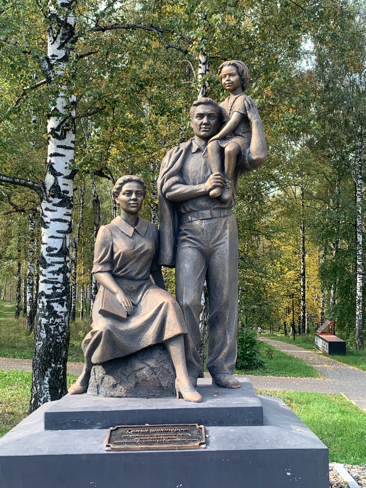 This is a bronze sculpture of a family in a park. The family consists of a father, mother, and child. The father stands in the center, holding a young child on his shoulders. The mother sits to the left of the father, holding a book. The sculpture is located in the city of Prokopyevsk, Russia, and is part of a memorial to the Shakhterov family. The park setting includes trees, grass, and a paved path.