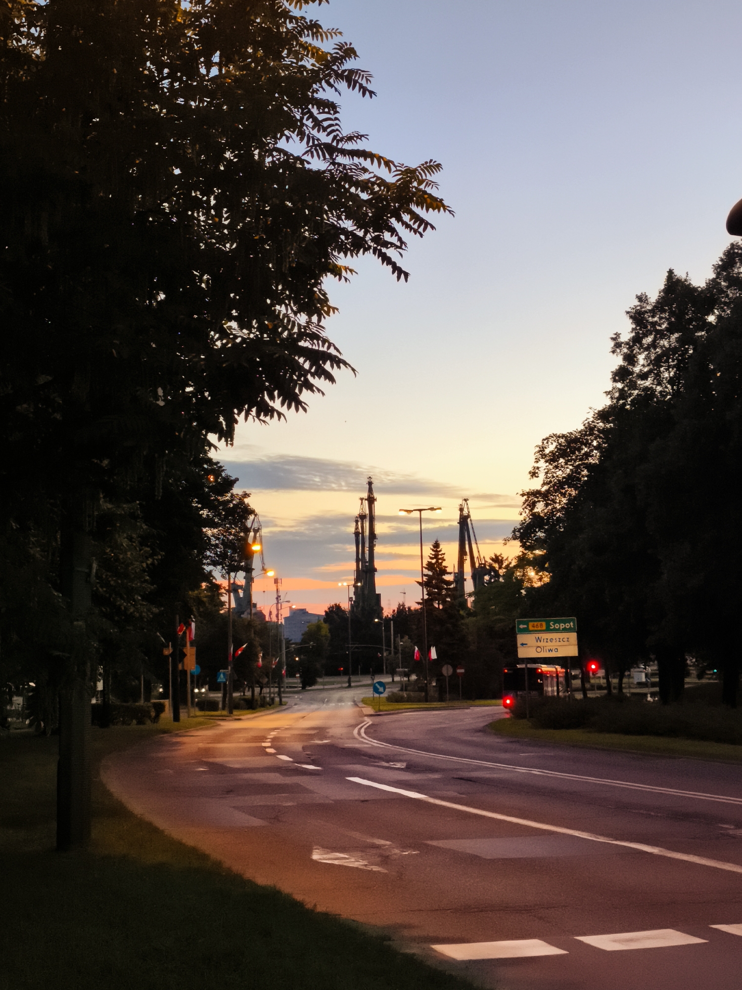 The image depicts a road leading towards a city skyline at dusk, with an orange and pink gradient in the sky. The foreground shows a curved asphalt road with white lane markings and a pedestrian crossing. To the left, a grassy embankment and the dark silhouette of a large tree are visible. On the right side of the road, a green road sign indicates directions to "Sopot" and "Wrzeszcz Oliwa." Streetlights and traffic lights are illuminated, casting a warm glow on the wet road surface. In the background, several tall structures, possibly cranes or industrial towers, rise above the trees. The overall scene suggests a quiet urban environment transitioning from day to night.