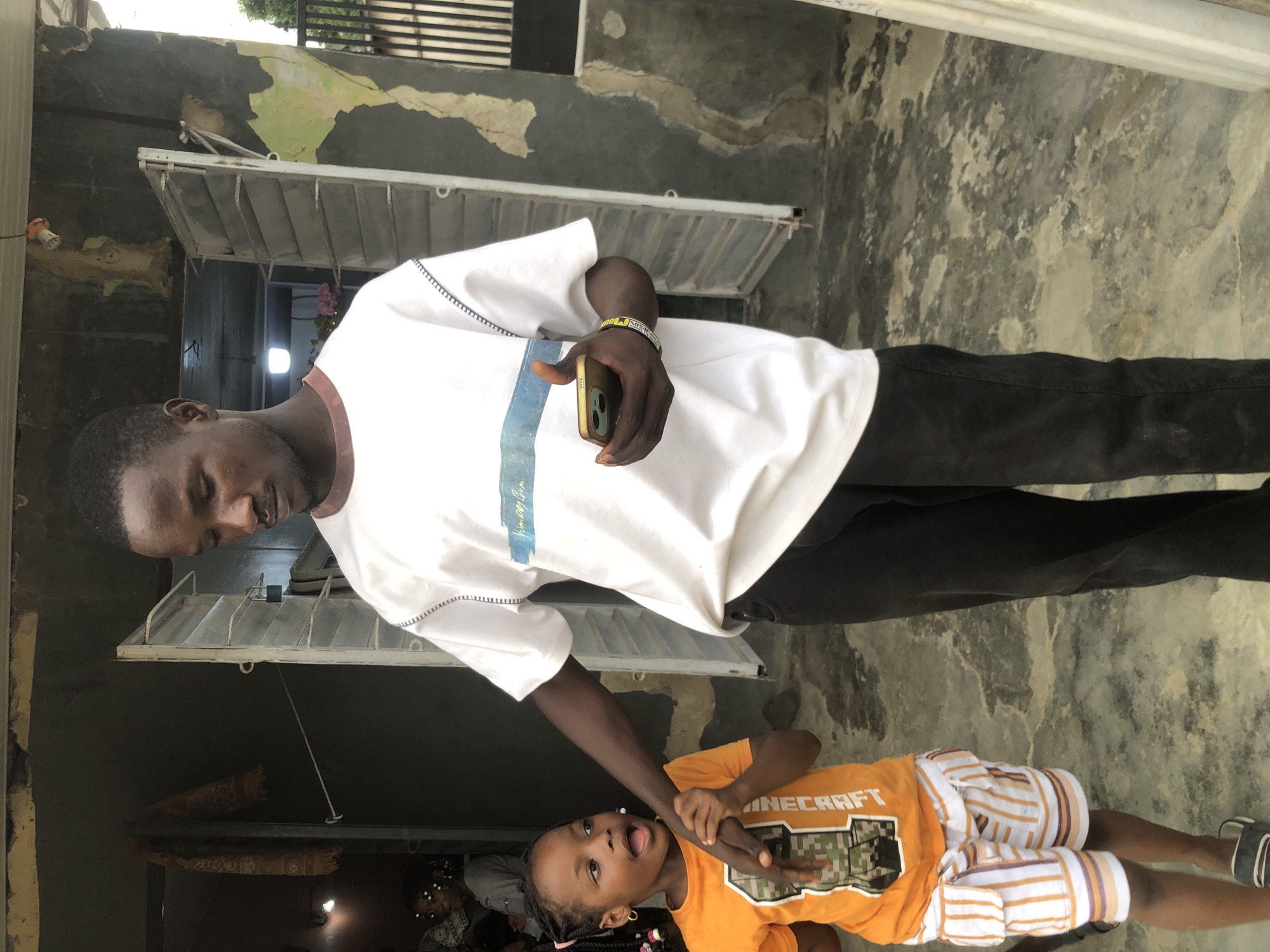 A young man and a young girl are standing outside a building in Ado, Nigeria. The man is wearing a white t-shirt and black pants, and he is holding a smartphone in his right hand. The girl is wearing an orange t-shirt and striped shorts, and she is holding the man's hand. The building behind them appears to be under construction or in disrepair, with peeling paint and exposed concrete. The ground is concrete and appears weathered. The lighting suggests it is daytime.