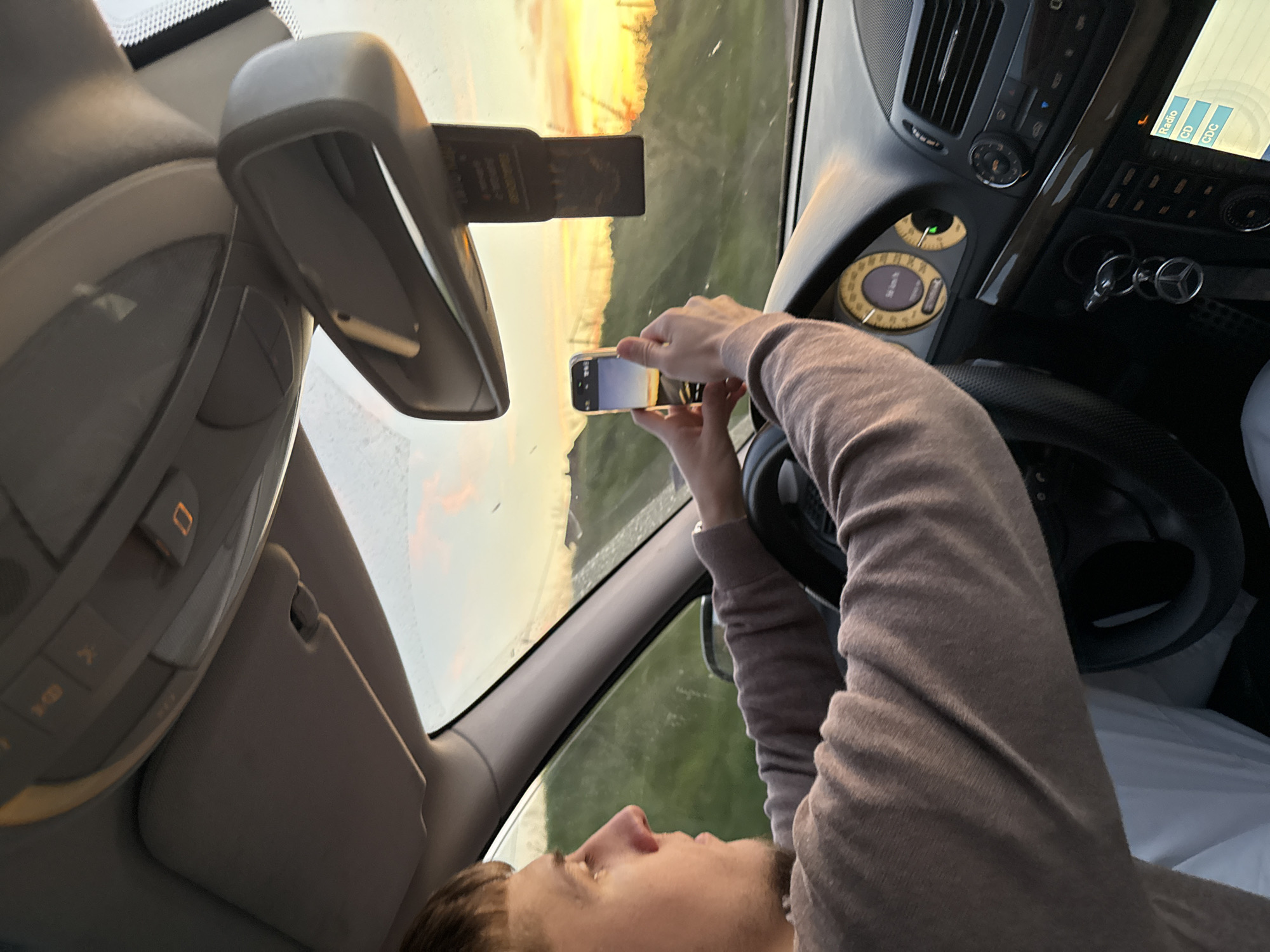 The image, taken inside a car, shows a person capturing the exterior view with a smartphone during what appears to be either sunrise or sunset. The perspective is from the passenger side, looking across the driver's seat towards the front windshield. The image is oriented sideways, with the car interior on the left and the outdoor scene on the right.
The primary subject is a person, likely male, with short, dark hair, seen in profile on the lower left. They are wearing a light brown or beige long-sleeved top. Their hands are holding a light-colored smartphone horizontally, aiming it through the windshield to photograph the vibrant sky. The phone's screen reflects the bright, orange and yellow sky, indicating it is actively recording or previewing a shot.
Inside the car, identifiable objects include the dashboard and controls of what appears to be a Mercedes-Benz, given the distinctive key in the ignition. The steering wheel is visible, as are circular gauges to its left, one showing "50 km/h" and a green indicator light. Above the steering wheel, part of the center console features air vents, climate control knobs, and a digital screen displaying "Radio", "CD", and "CDC". A dark wood-grain trim accentuates the dashboard. The rearview mirror is in the upper left, with a dark, rectangular attachment. An overhead console is partially visible, showing buttons and an illuminated orange square indicator with a "0" symbol.
Outside the car, the sky is dominated by warm, golden, and orange hues, characteristic of early morning or late afternoon, suggesting clear weather. Below the horizon, a green landscape, possibly fields or scattered trees, is visible, along with faint outlines of what could be utility poles or fences. The car appears to be in motion, or has recently been, given the "50 km/h" reading. The setting is in Sofia, Bulgaria.