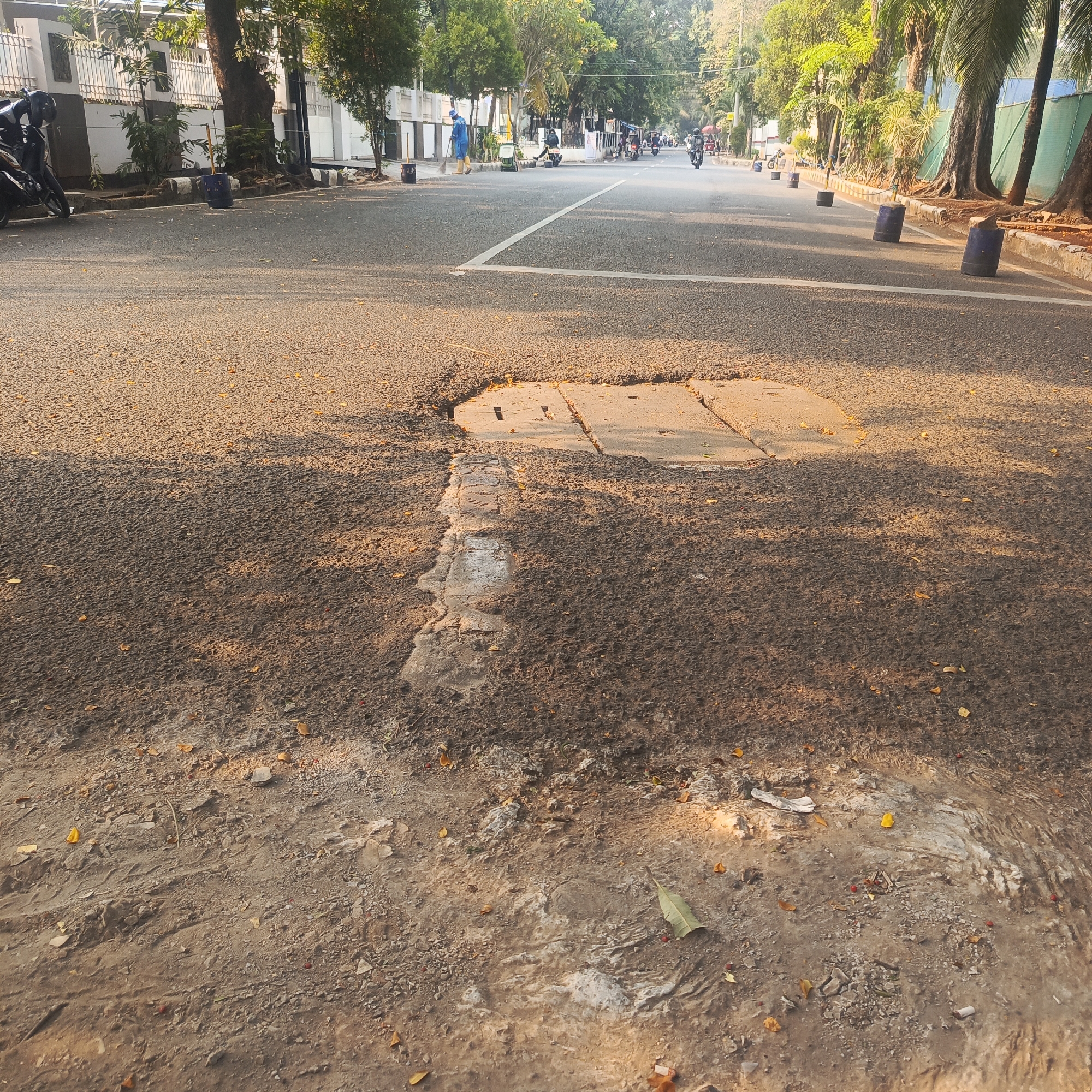 The image captures a street scene in South Jakarta, Indonesia, during daylight hours. The perspective is low, looking forward along a paved road with a prominent white line dividing the lanes. The road surface is asphalt, showing signs of wear and tear, particularly a large pothole in the foreground with a concrete slab partially covering it. The shoulders of the road are unpaved, consisting of dirt and debris, with scattered leaves and small red berries.
On the left side of the road, a white building with decorative fencing is visible, along with trees and a parked motorcycle with a black helmet on its seat. In the distance, several motorcycles and a person in a blue uniform with a broom are seen on the road, suggesting some form of street cleaning or maintenance activity.
The right side of the road features a sidewalk with palm trees and other vegetation. Several blue cylindrical objects, possibly traffic cones or barriers, are placed along the edge of the road. The sunlight casts long shadows across the scene, indicating it is either early morning or late afternoon. The overall impression is a typical urban street environment in South Jakarta, showing signs of both daily activity and the need for road upkeep.