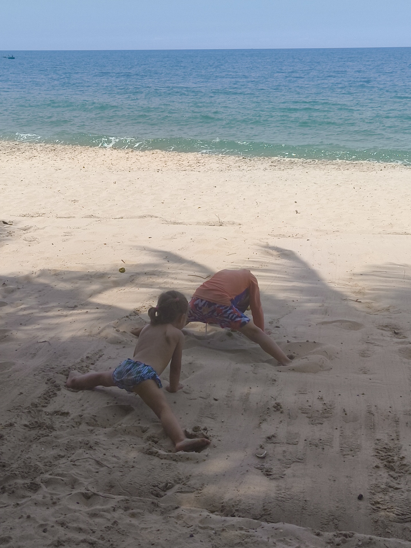 Two children are on a sandy beach in front of the ocean. The child on the left is a young girl with her back to the camera, wearing blue and green patterned swim shorts. She is in a kneeling split position with her legs extended to the sides. The child on the right is wearing an orange long-sleeved shirt and patterned shorts, and is in a crawling or squatting position with their head facing downwards into the sand. The sand is light brown and textured with footprints and shadows cast by palm trees. The ocean is blue with small waves and a clear sky above. In the far distance, a small boat is visible on the horizon.