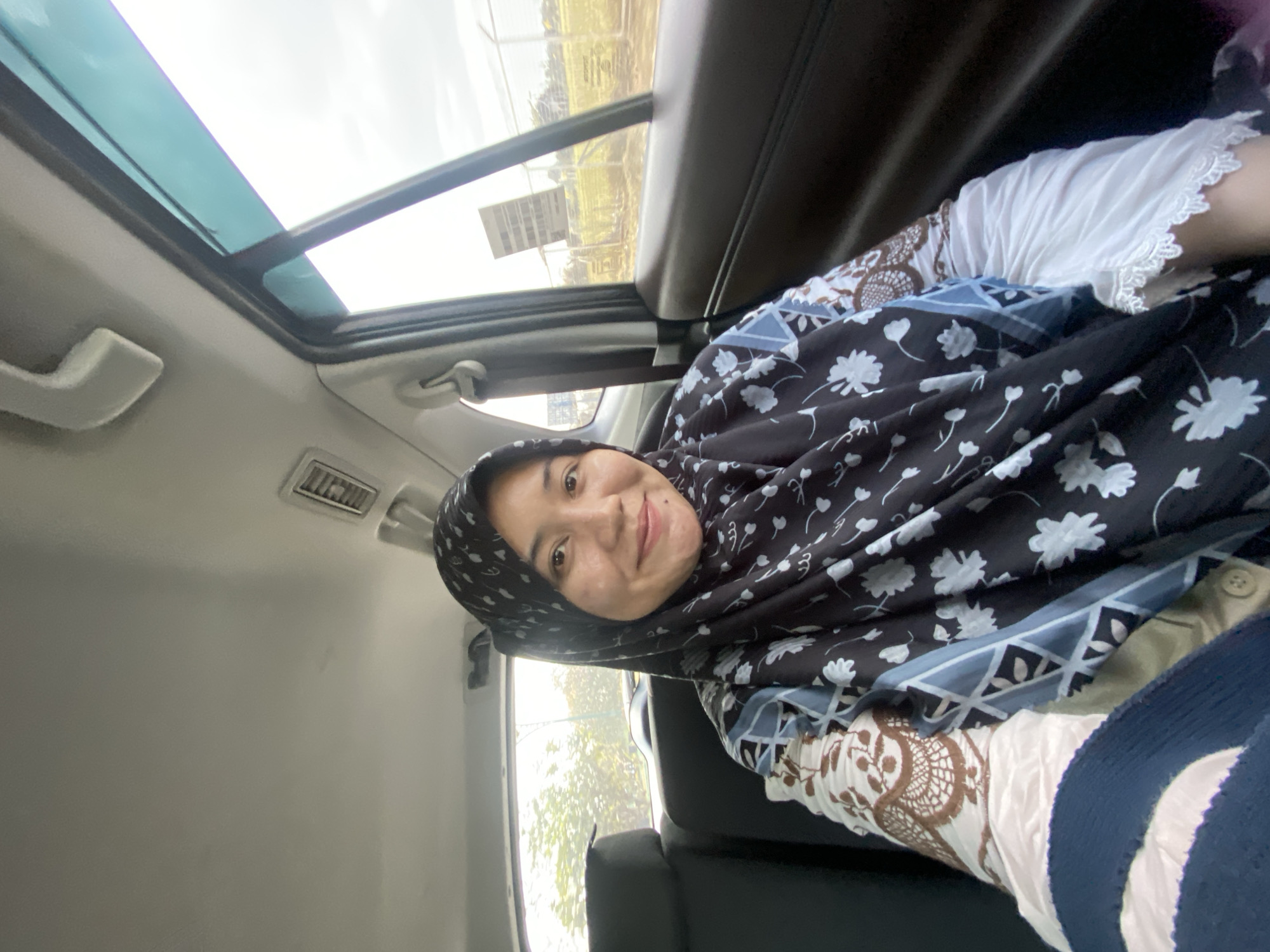 A woman wearing a hijab and a patterned shirt sits in the back seat of a car. She is smiling at the camera. The car interior is visible, including the seats, ceiling, and window. Outside the window, trees and a fence can be seen. The image was taken in Banjarbaru, Indonesia.