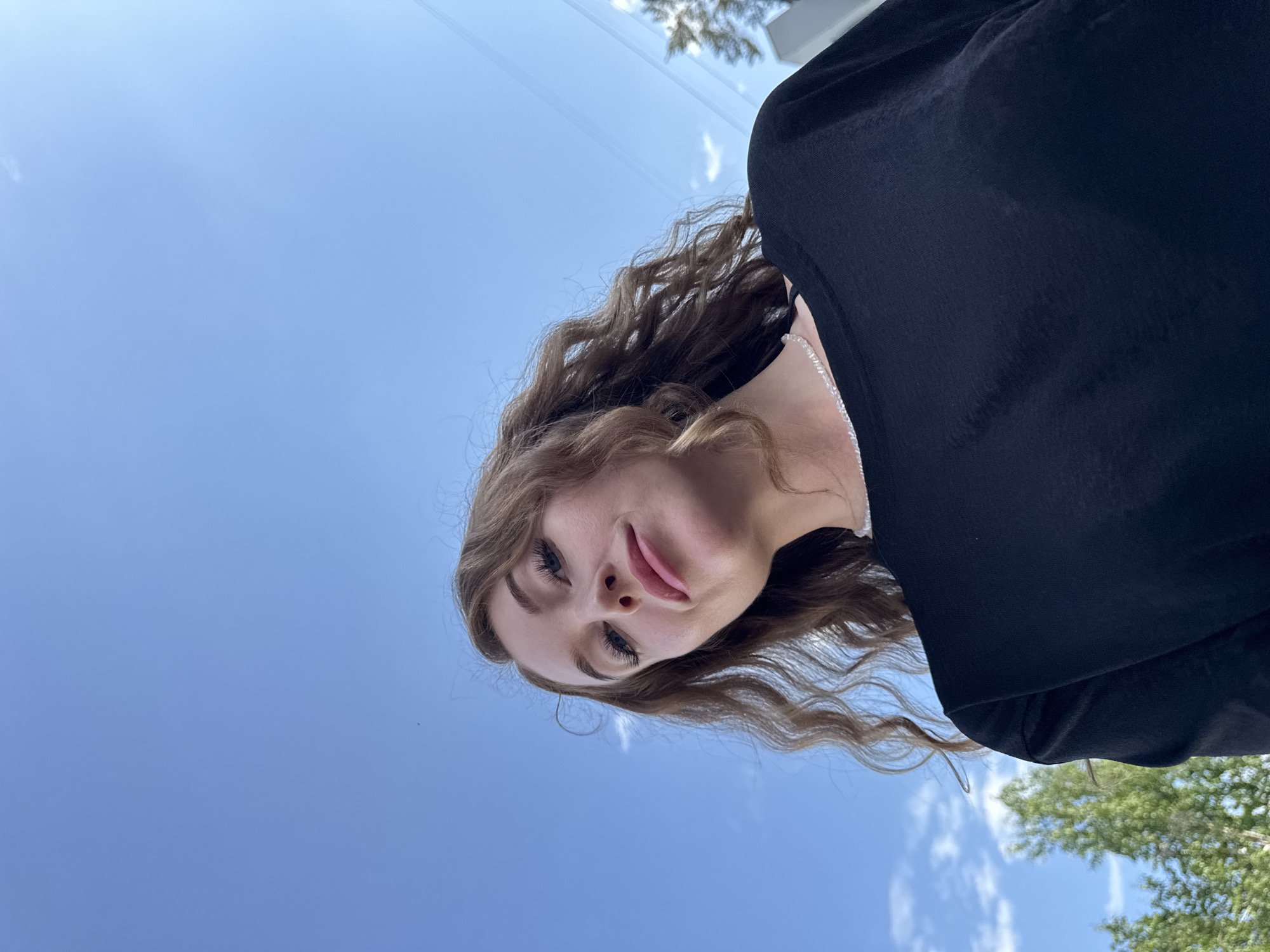 A young woman with long, wavy light brown hair is looking down at the camera. She is wearing a black top and a thin silver necklace. The background is a bright blue sky with a few scattered clouds and a glimpse of green trees in the bottom right corner. The location is unknown.