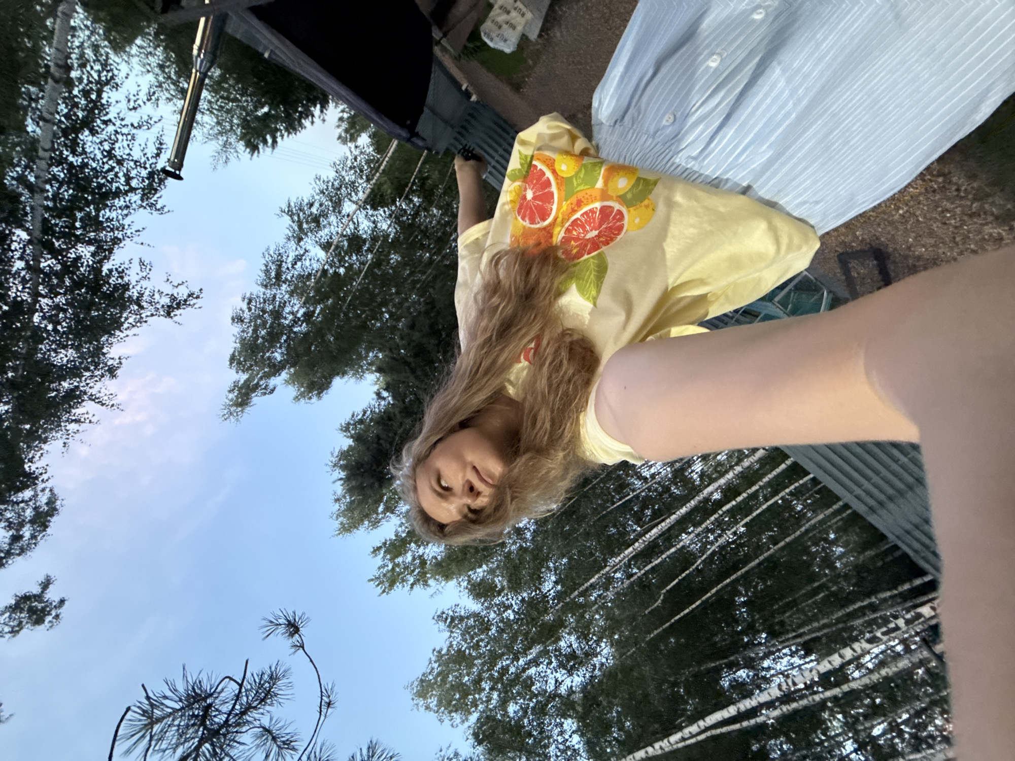 A young woman with blonde hair, wearing a yellow t-shirt with a grapefruit design and a light blue striped skirt, is captured in a low-angle selfie. She is outdoors, with a backdrop of tall trees, some appearing to be birch trees with white bark, and a clear sky with faint clouds. To the right, part of a dark-colored structure with a metal pipe extending upwards is visible, along with a glimpse of a gravel surface and possibly some distant structures. The woman is looking up and to the side, with her arm extended holding the camera. The setting appears to be a natural or semi-rural environment, possibly on the outskirts of Prokudino, Russia, given the typical vegetation and outdoor structures.